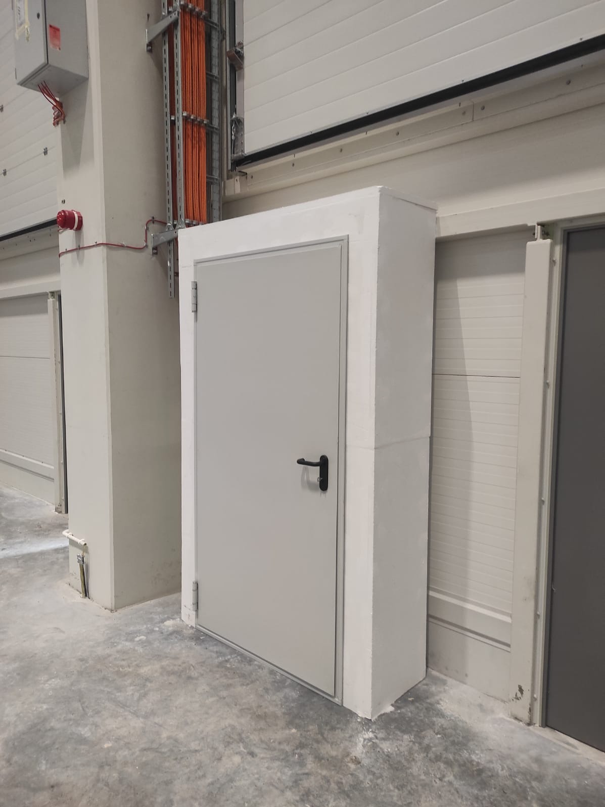 Szara, metalowa, przeciwpożarowa brama w betonowej obudowie, zamontowana w hali przemysłowej. Nad bramą widoczne kable w korytkach. W tle brama garażowa.