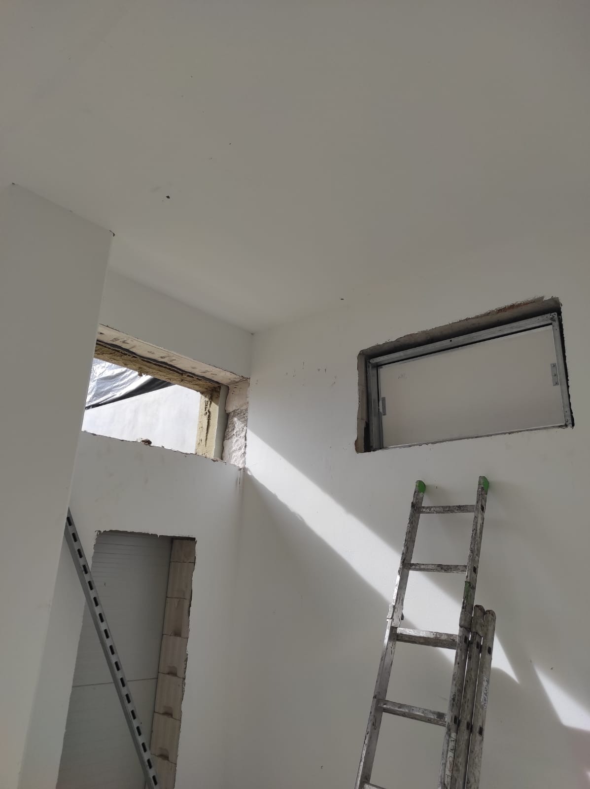 Wnętrze w trakcie prac murarskich: otwory okienne w surowym stanie, drabina oparta o ścianę. Widoczne ślady po kuciu i przygotowaniu do montażu okien.