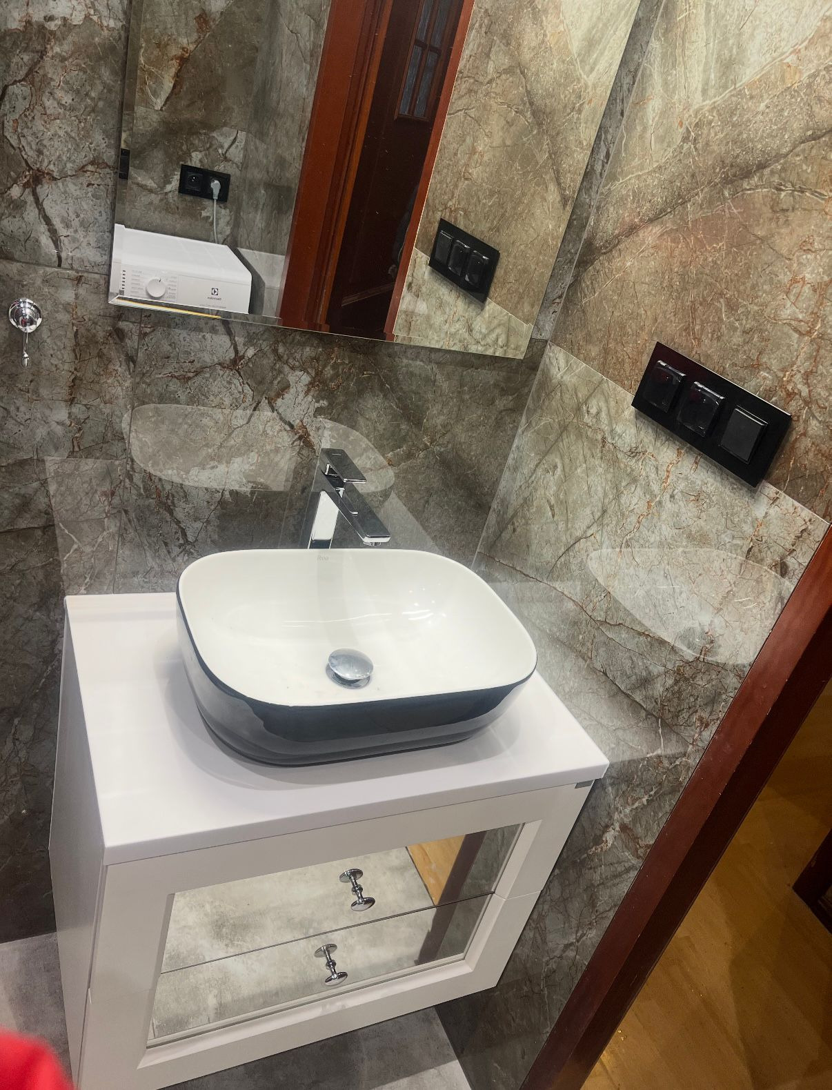 Nowoczesna umywalka nablatowa w łazience z marmurowymi płytkami i lustrem, w którym odbija się pralka. Biała szafka pod umywalką z lustrzanymi frontami.