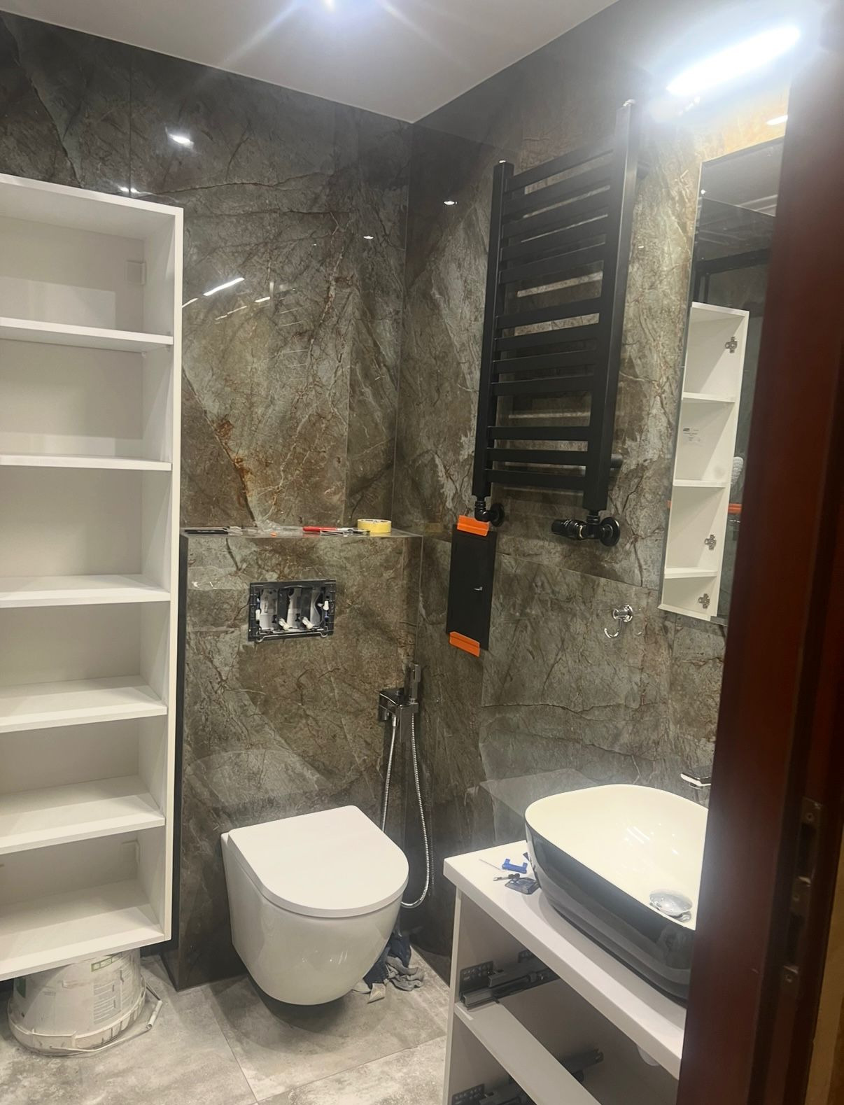 Wnętrze łazienki w trakcie wykańczania: marmurowe płytki, biała toaleta podtynkowa, czarny grzejnik drabinkowy, biała szafka z umywalką i regał. Widoczne narzędzia.