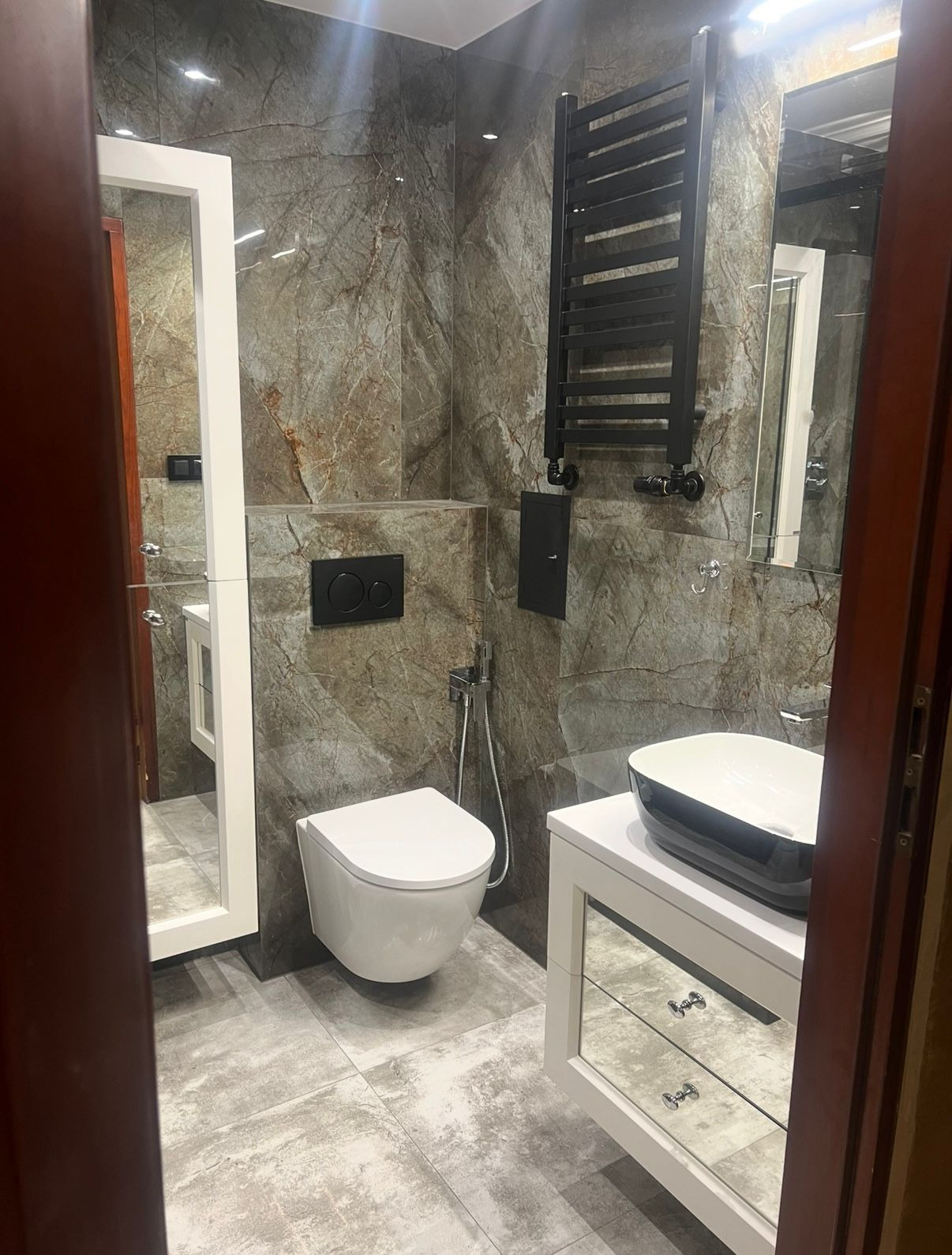 Elegancka łazienka z marmurowymi płytkami, wiszącą toaletą, grzejnikiem drabinkowym i umywalką nablatową na szafce z lustrzanymi frontami. Nowoczesny design.