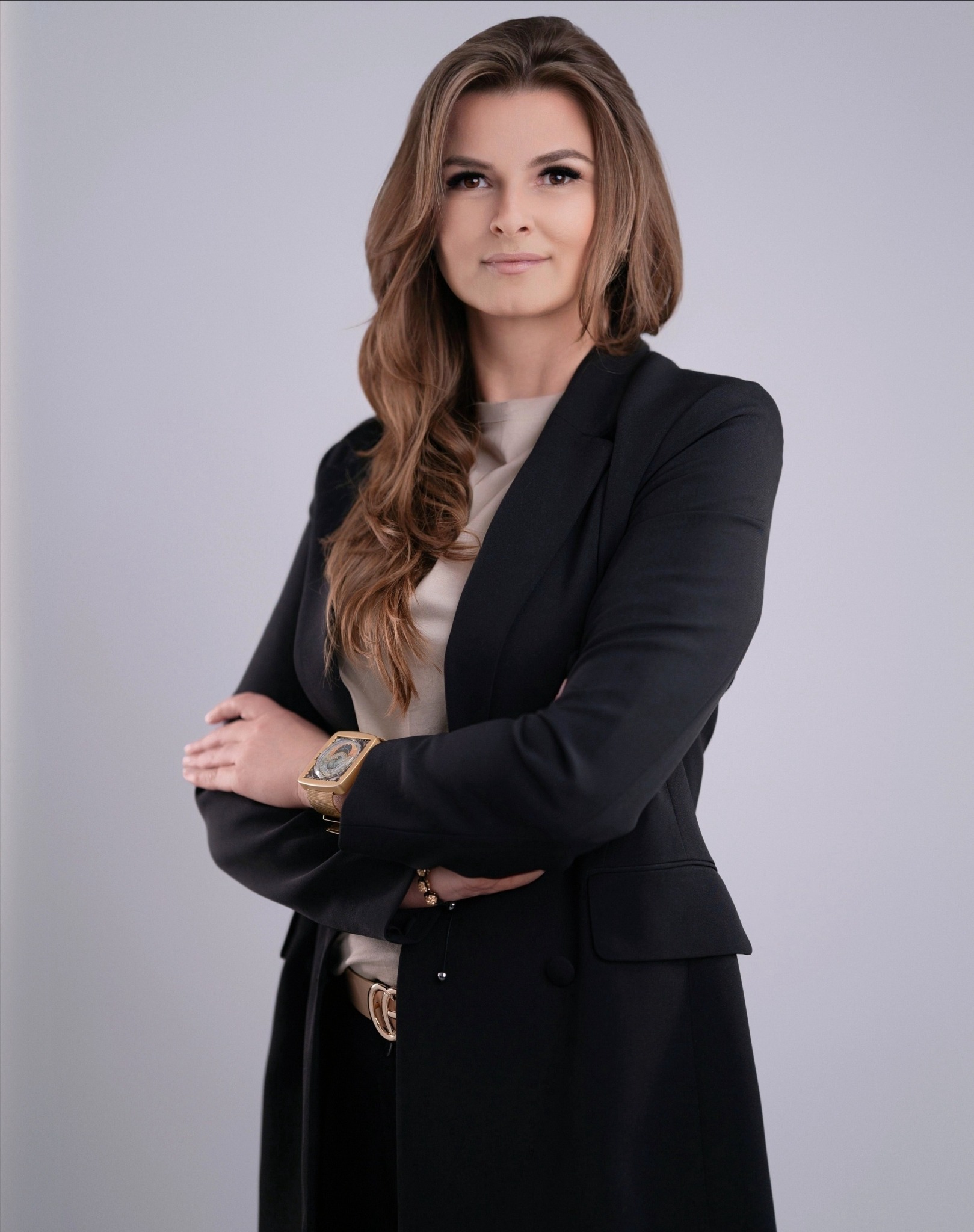 Profesjonalny portret kobiety w czarnej marynarce, ze złotym zegarkiem i paskiem, na jasnoszarym tle. Uśmiechnięta agentka nieruchomości.