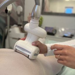 Marta Buchar COSMETOLOGY - Urządzenie do modelowania sylwetki w trakcie zabiegu na ciele pacjenta ubranego w specjalny kombinezon. W tle widoczne lustro i inne urządzenia.