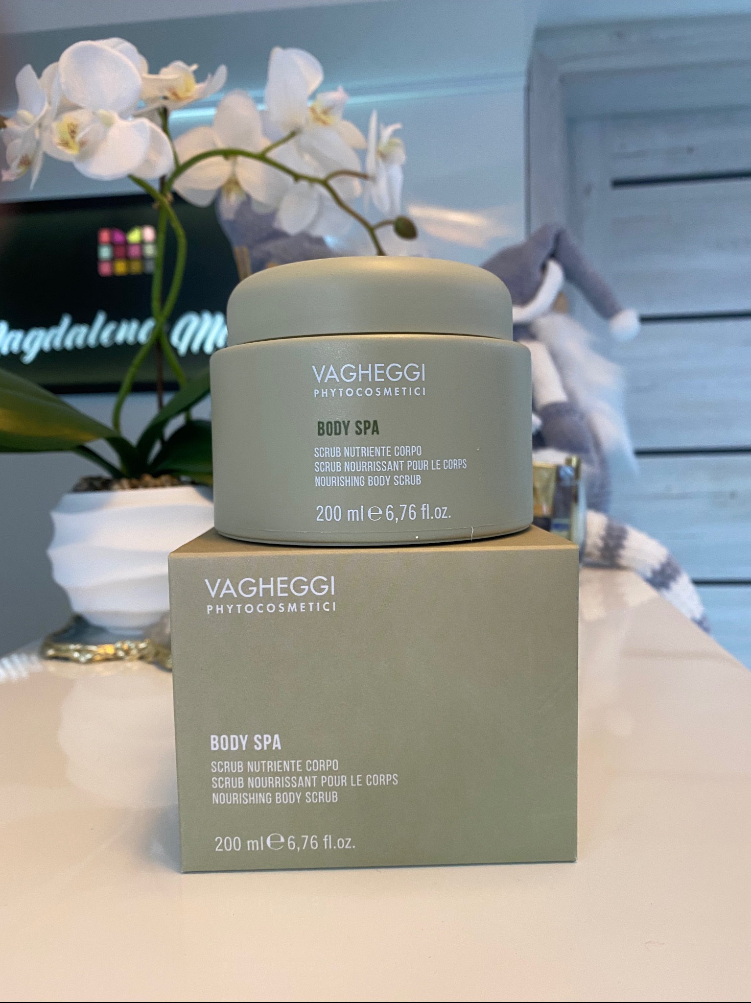 Zestaw kosmetyków Vagheggi Body Spa: peeling do ciała w minimalistycznym opakowaniu, z kwiatem orchidei w tle, na jasnym blacie. Elegancki i subtelny.