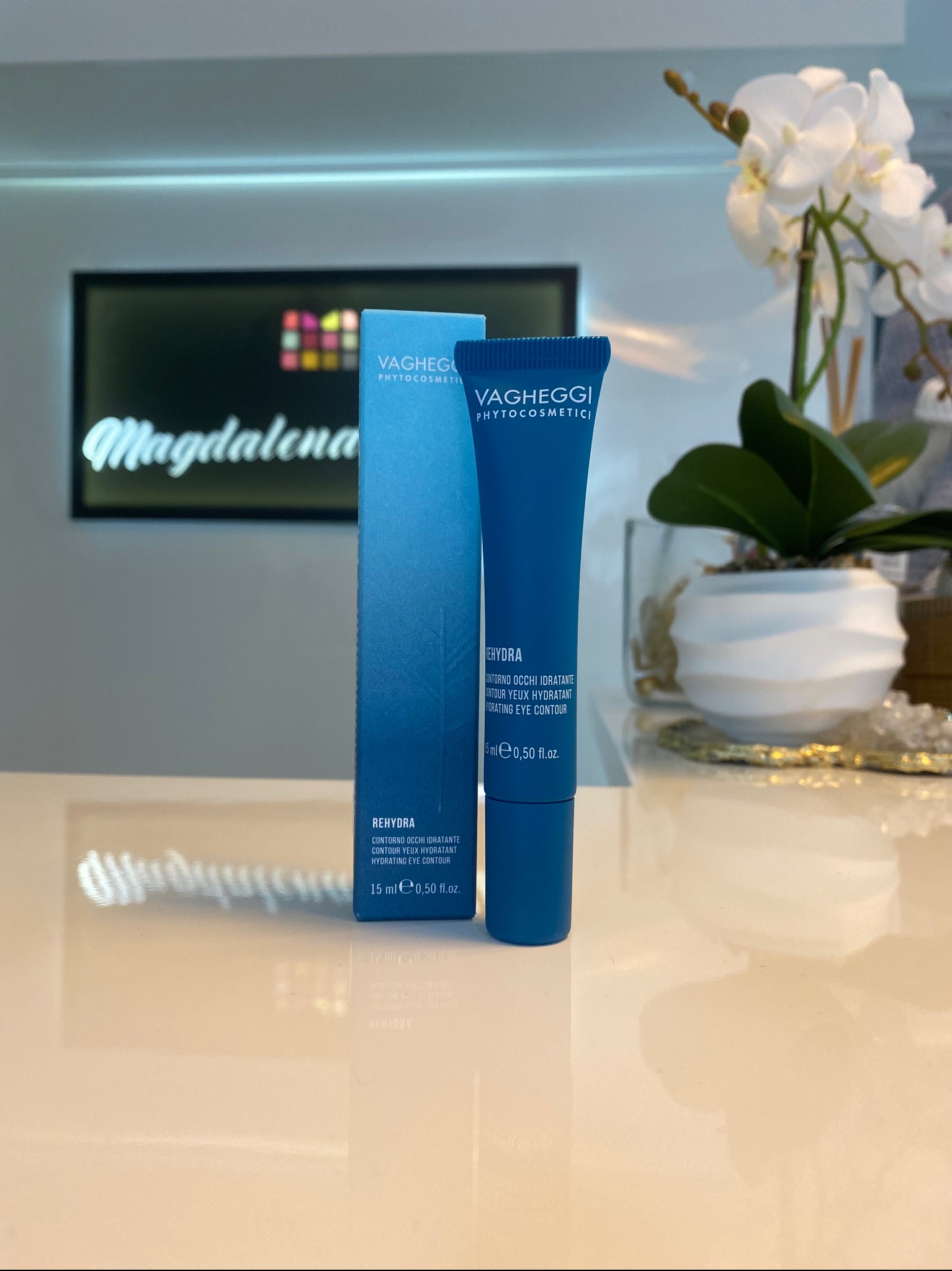 Niebieski zestaw kosmetyków Vagheggi Rehydra do pielęgnacji okolic oczu, opakowanie i tubka, na błyszczącym blacie z odbiciem, w tle kwiaty i logo salonu.
