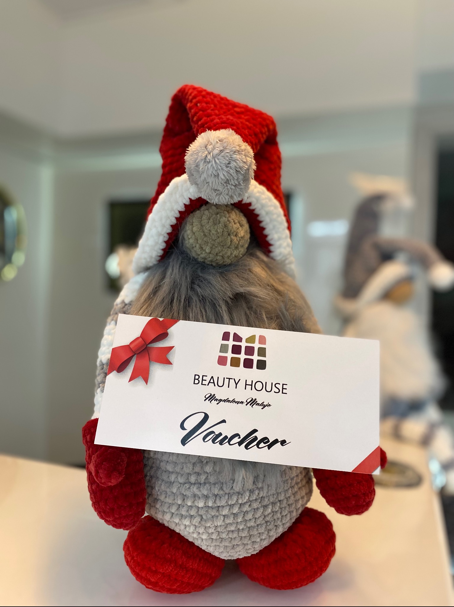 Ręcznie robiony krasnal trzymający voucher do Beauty House. Czerwony kapelusz, szara broda, białe tło. Prezent świąteczny na zabiegi kosmetyczne.
