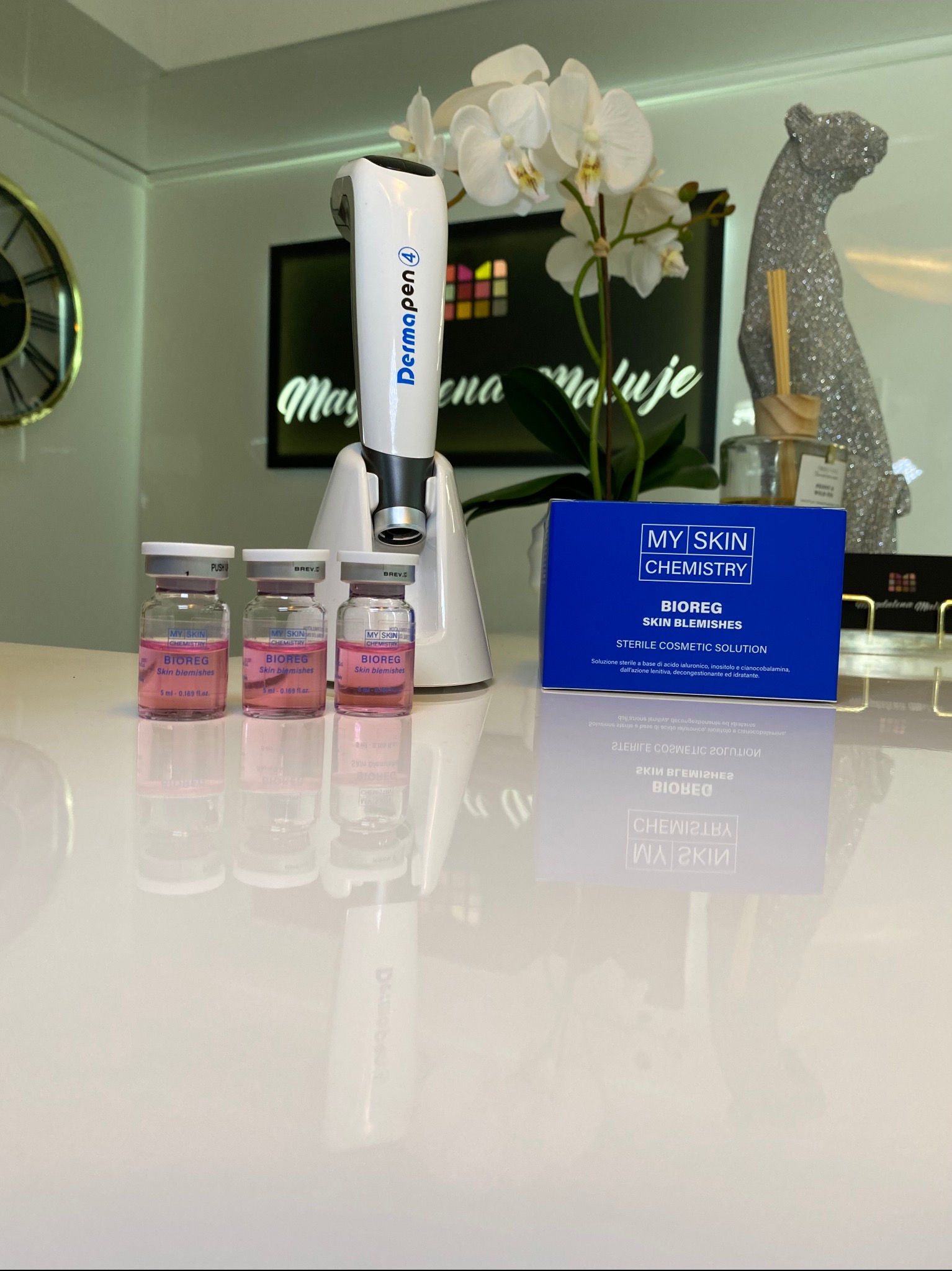 DermaPen 4 z ampułkami Bioreg Skin Blemishes i pudełkiem My Skin Chemistry na błyszczącym blacie, w tle kwiaty i dekoracja pantery, sugerujące zabieg kosmetyczny.