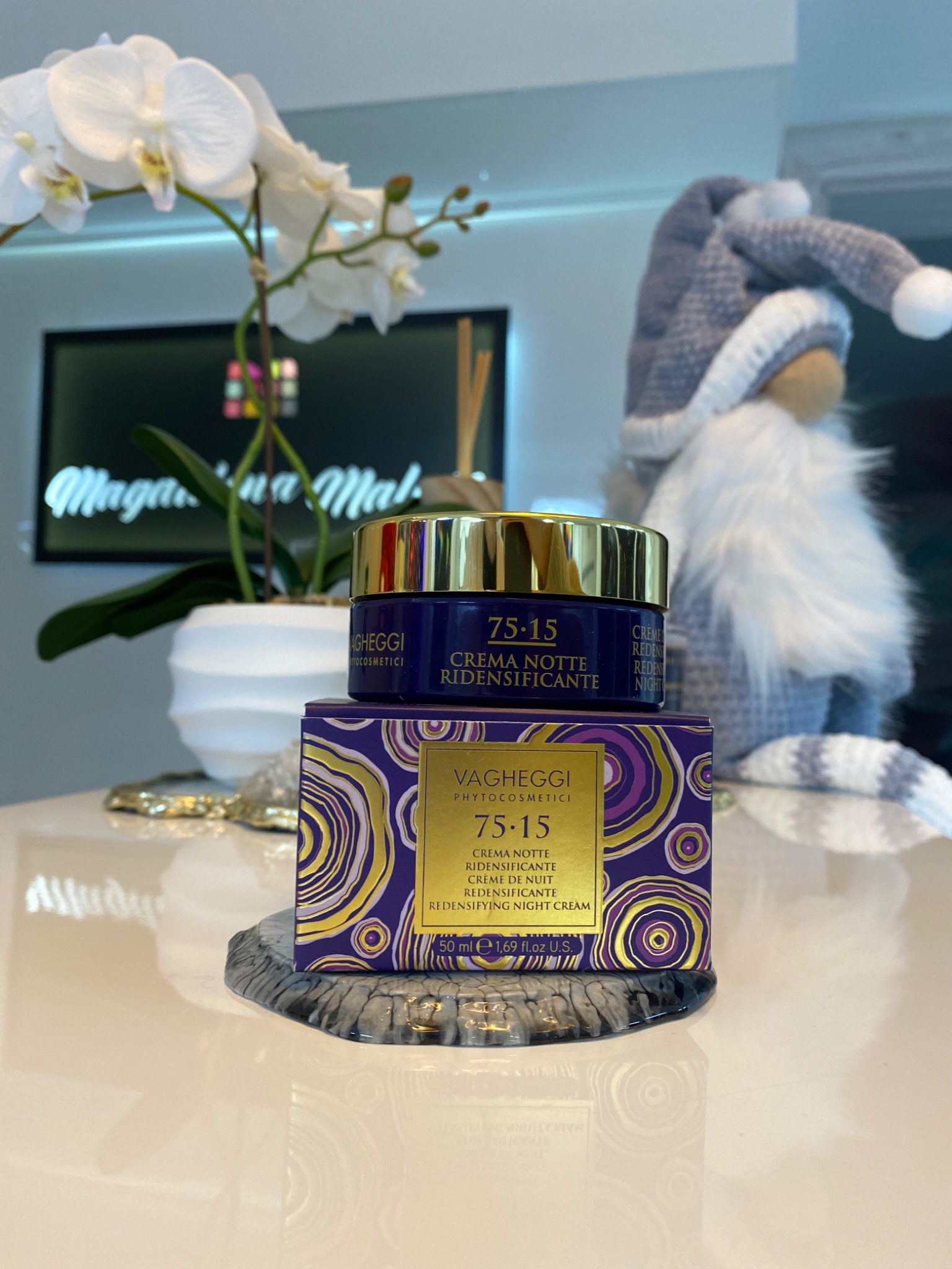 Fioletowy krem na noc Vagheggi 75.15 na pudełku, z kwiatami i figurką w tle. Produkt do pielęgnacji skóry w salonie kosmetycznym.