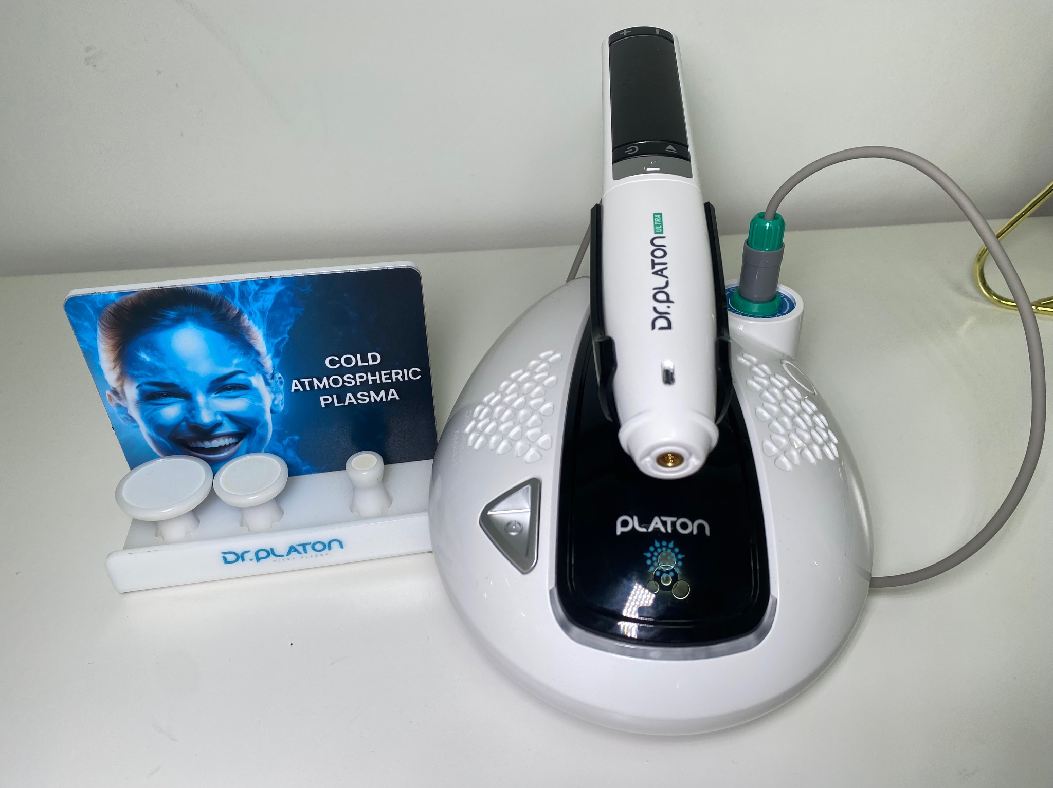 Urządzenie Dr. Platon Ultra do zabiegów Cold Atmospheric Plasma, z aplikatorem i podstawką, obok ekspozytor z wizerunkiem kobiety i akcesoriami.