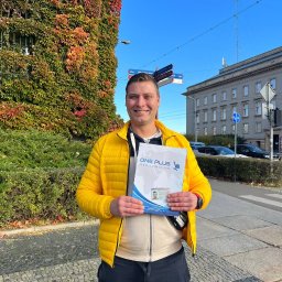 ONE PLUS Sp. z o.o. - Uśmiechnięty mężczyzna w żółtej kurtce prezentuje folder 'ONE PLUS HR & Relocation Solutions' na tle budynku z bluszczem i drogowskazów.