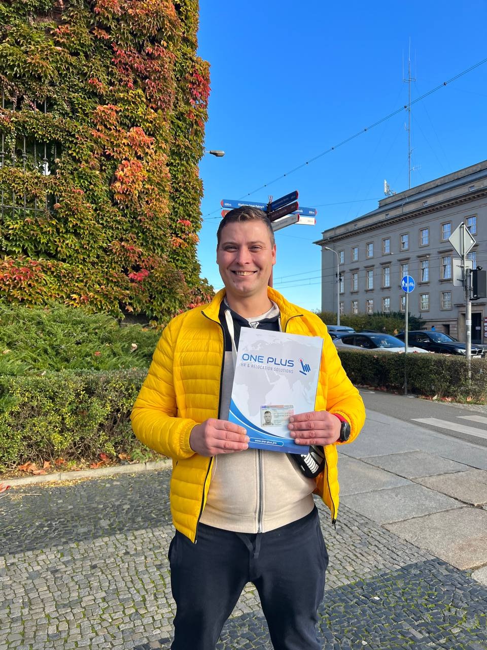 Uśmiechnięty mężczyzna w żółtej kurtce prezentuje folder 'ONE PLUS HR & Relocation Solutions' na tle budynku z bluszczem i drogowskazów.
