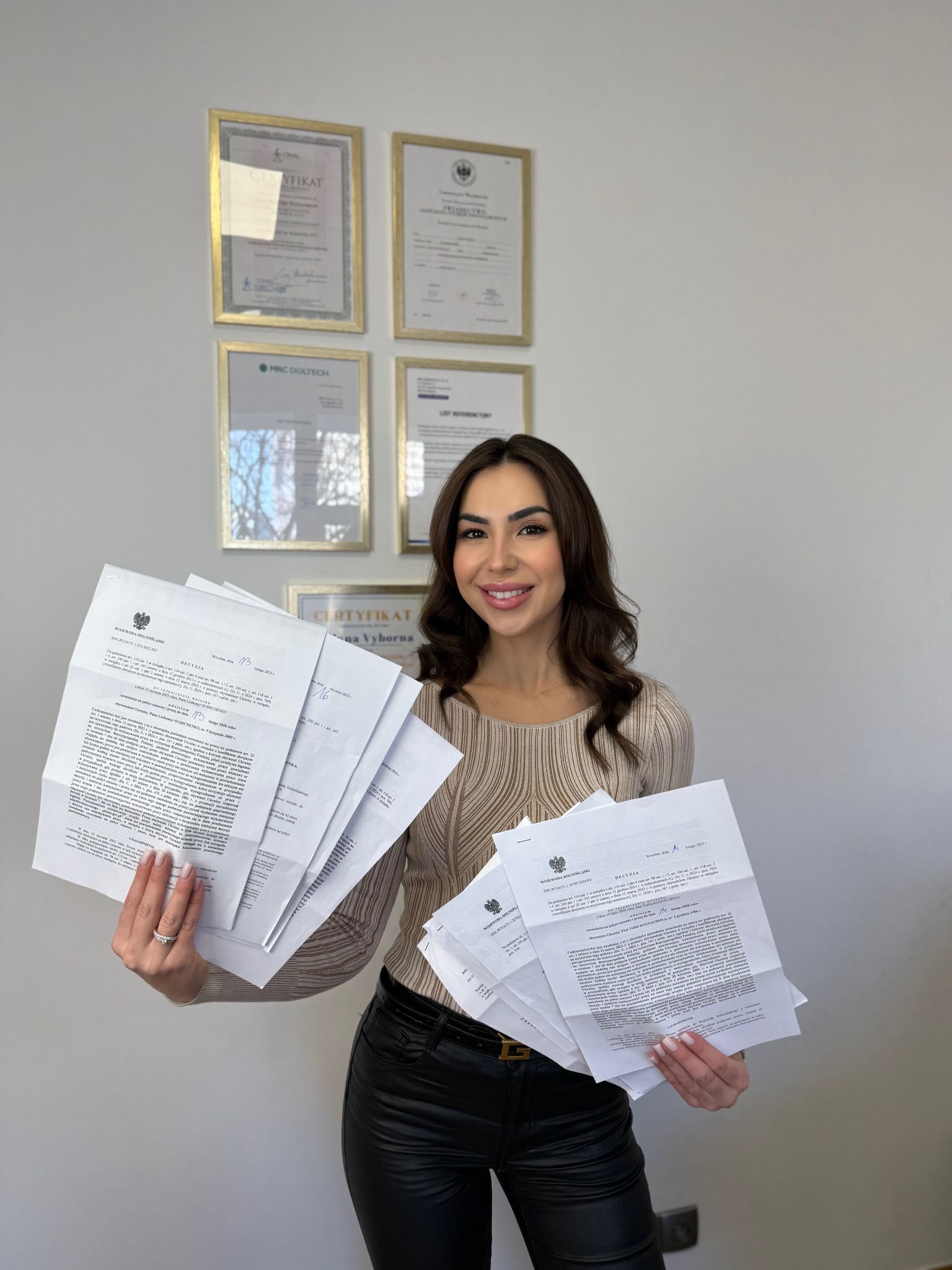 Uśmiechnięta kobieta trzymająca plik dokumentów urzędowych na tle ściany z certyfikatami. Widoczne decyzje administracyjne z godłem Polski.
