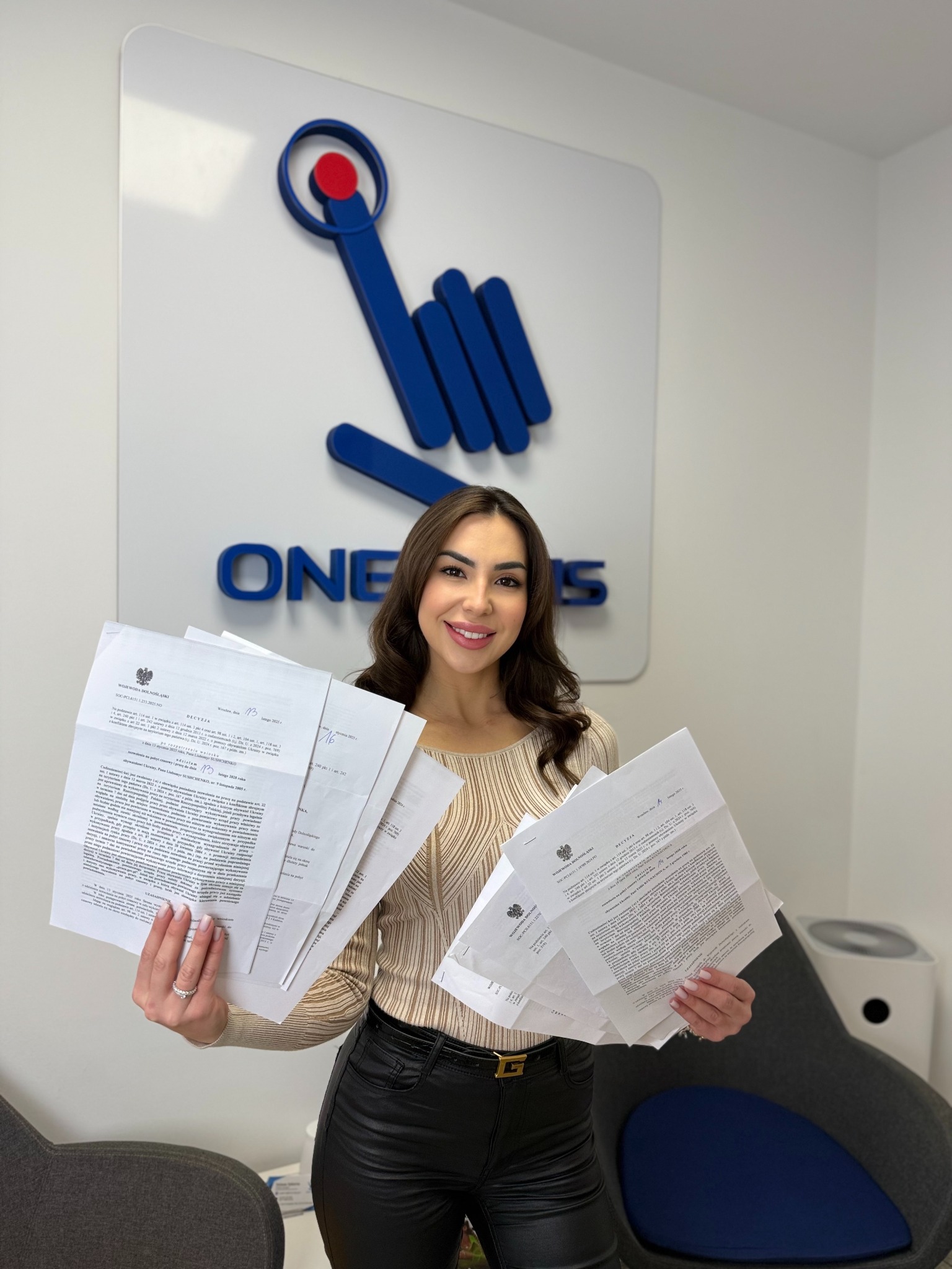 Uśmiechnięta kobieta trzyma plik dokumentów z pieczęciami na tle biura z logo firmy. Dokumenty urzędowe, prawdopodobnie decyzje administracyjne.