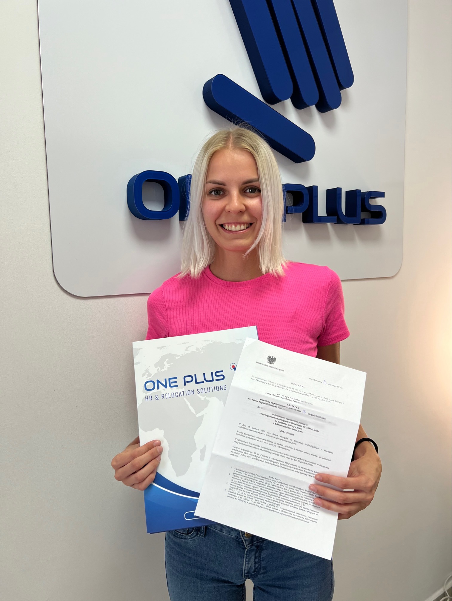 Kobieta z blond włosami prezentuje dokumenty firmy One Plus HR & Relocation Solutions, stojąc na tle logo firmy. Widoczna decyzja administracyjna.