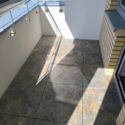 Perfekcyjne Wykończenia Marek Nowakowski - Wykończony balkon z szarymi płytkami imitującymi kamień, z metalową barierką i białymi ścianami. Widoczne refleksy słoneczne na podłodze.