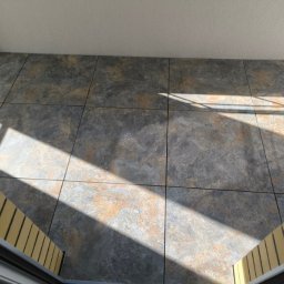 Perfekcyjne Wykończenia Marek Nowakowski - Ułożona podłoga z płytek w odcieniach szarości i beżu, oświetlona promieniami słonecznymi wpadającymi przez okno. Fragment schodów w prawym dolnym rogu.