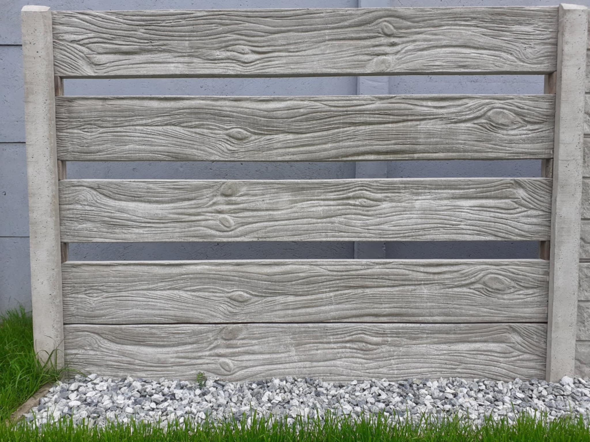 Betonowe przęsła ogrodzeniowe imitujące drewno, z widoczną fakturą słojów, osadzone na betonowych słupkach. U podstawy trawa i szare kamienie.