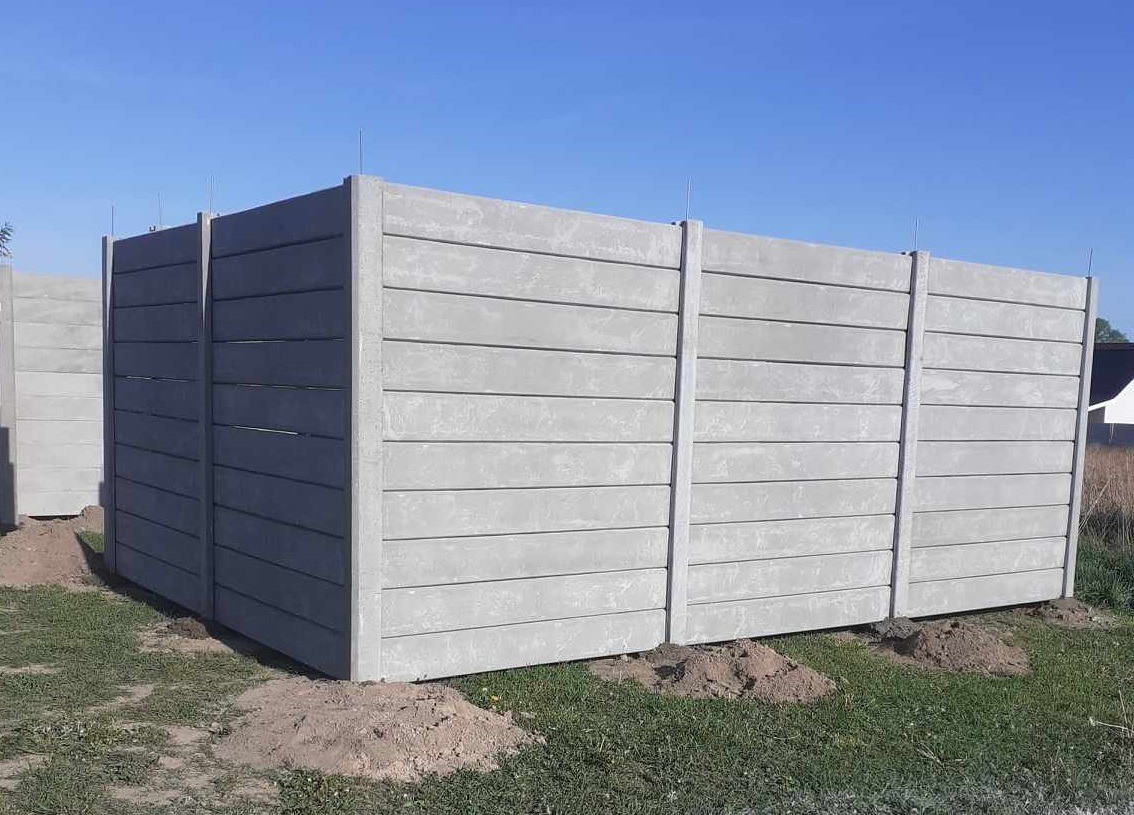 Szary, prefabrykowany płot betonowy z poziomymi panelami, świeżo zamontowany na trawiastym terenie z kopcami ziemi u podstawy, pod błękitnym niebem.