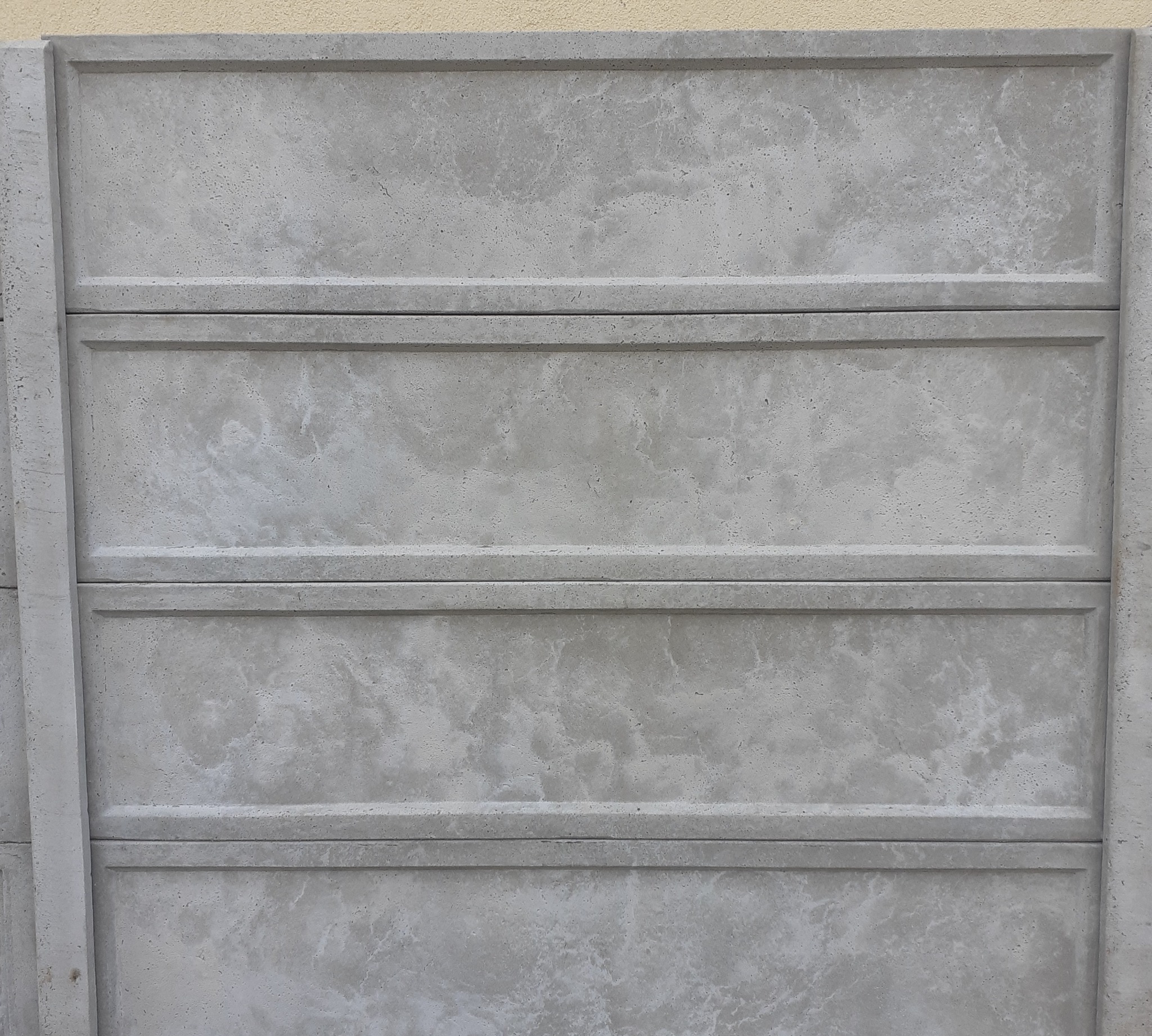 Fragment szarego, prefabrykowanego ogrodzenia betonowego z widoczną fakturą betonu i elementami konstrukcyjnymi. Tło stanowi jasna ściana.