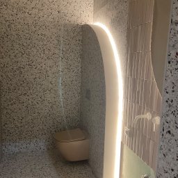 Drywall Daniel Twardowski - Wykończona łazienka z toaletą w kolorze beżowym, ściany i podłoga wyłożone terazzo, podświetlona wnęka z ledami, pionowe płytki na ścianie.