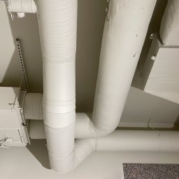 Drywall Daniel Twardowski - Białe rury wentylacyjne i klimatyzacyjne pod sufitem, widok z dołu. Elementy instalacji pomalowane na biało, widoczne łączenia i mocowania.