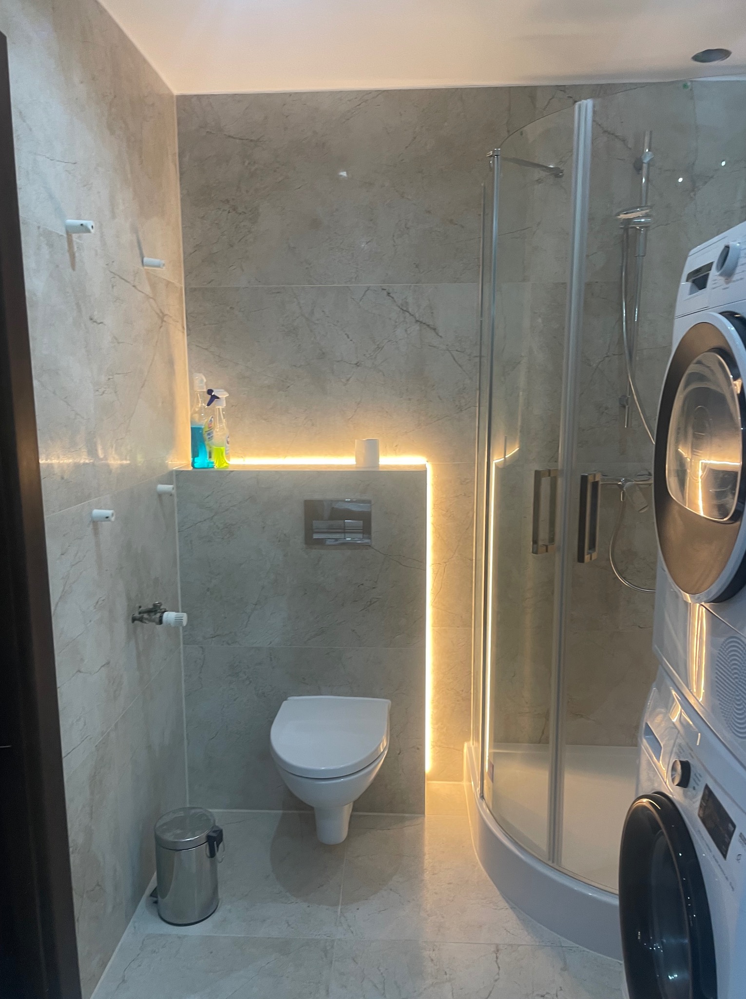 Wykończona łazienka w beżowych płytkach z podświetleniem LED, kabiną prysznicową i pralką. Widoczny kosz na śmieci i toaleta. Minimalistyczny design.