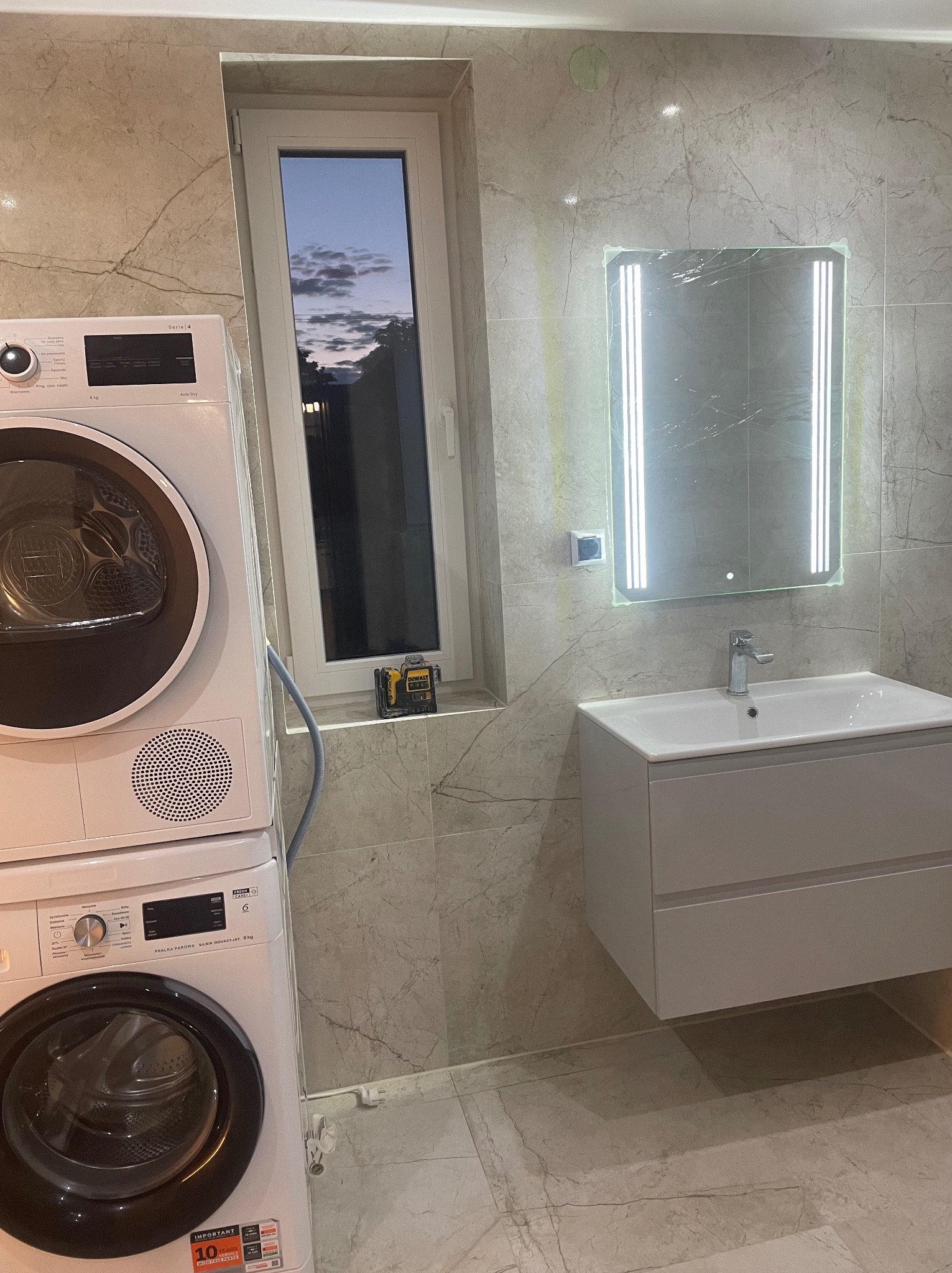 Wykończona łazienka w beżowych płytkach imitujących marmur. Umywalka z szafką, podświetlane lustro, pralka i suszarka w kolumnie. Poziomica laserowa na parapecie.