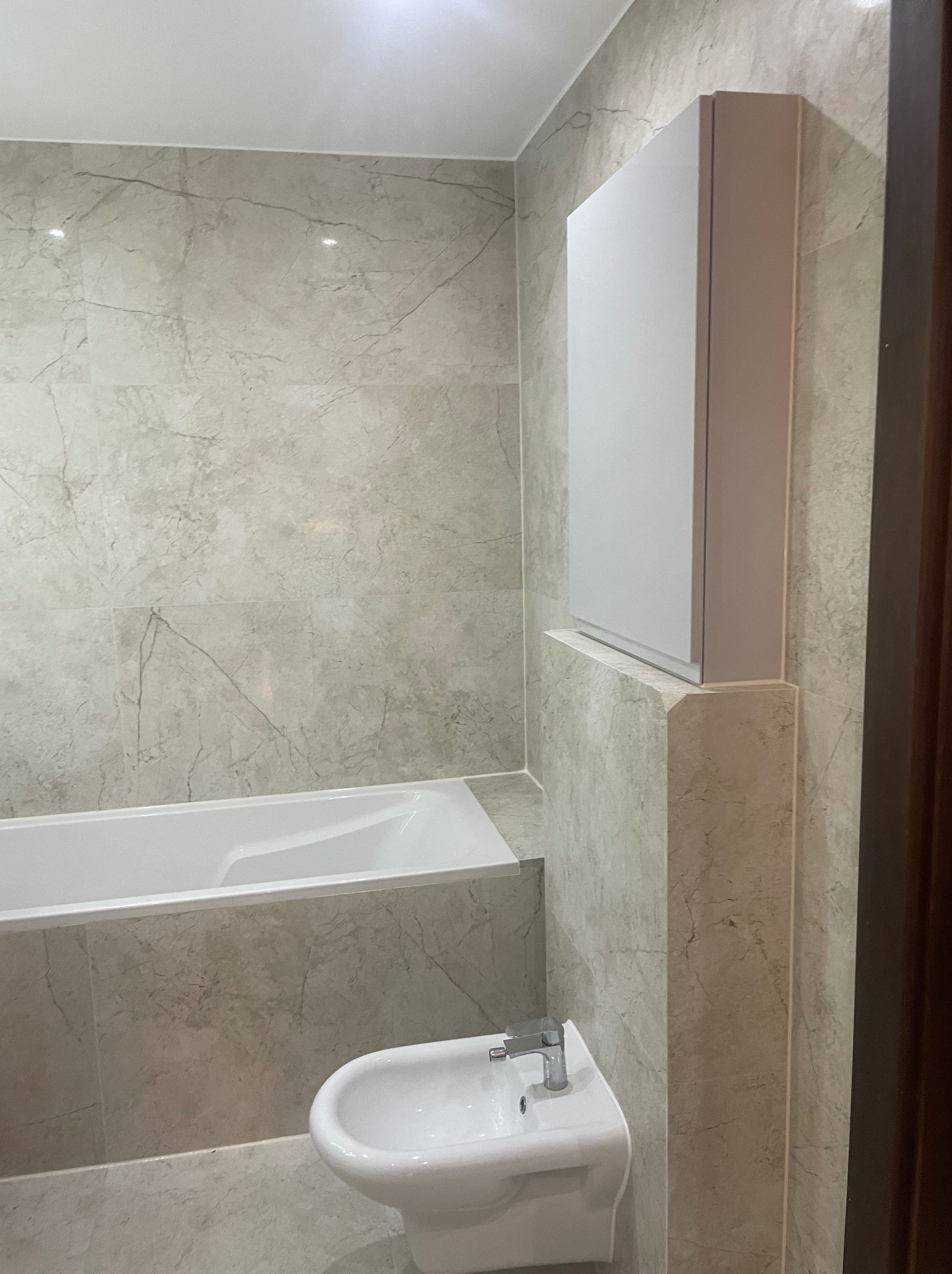 Wykończona łazienka z wanną, bidetem i szafką, wyłożona płytkami imitującymi marmur. Ujęcie z perspektywy narożnika, widoczne detale armatury i oświetlenia.