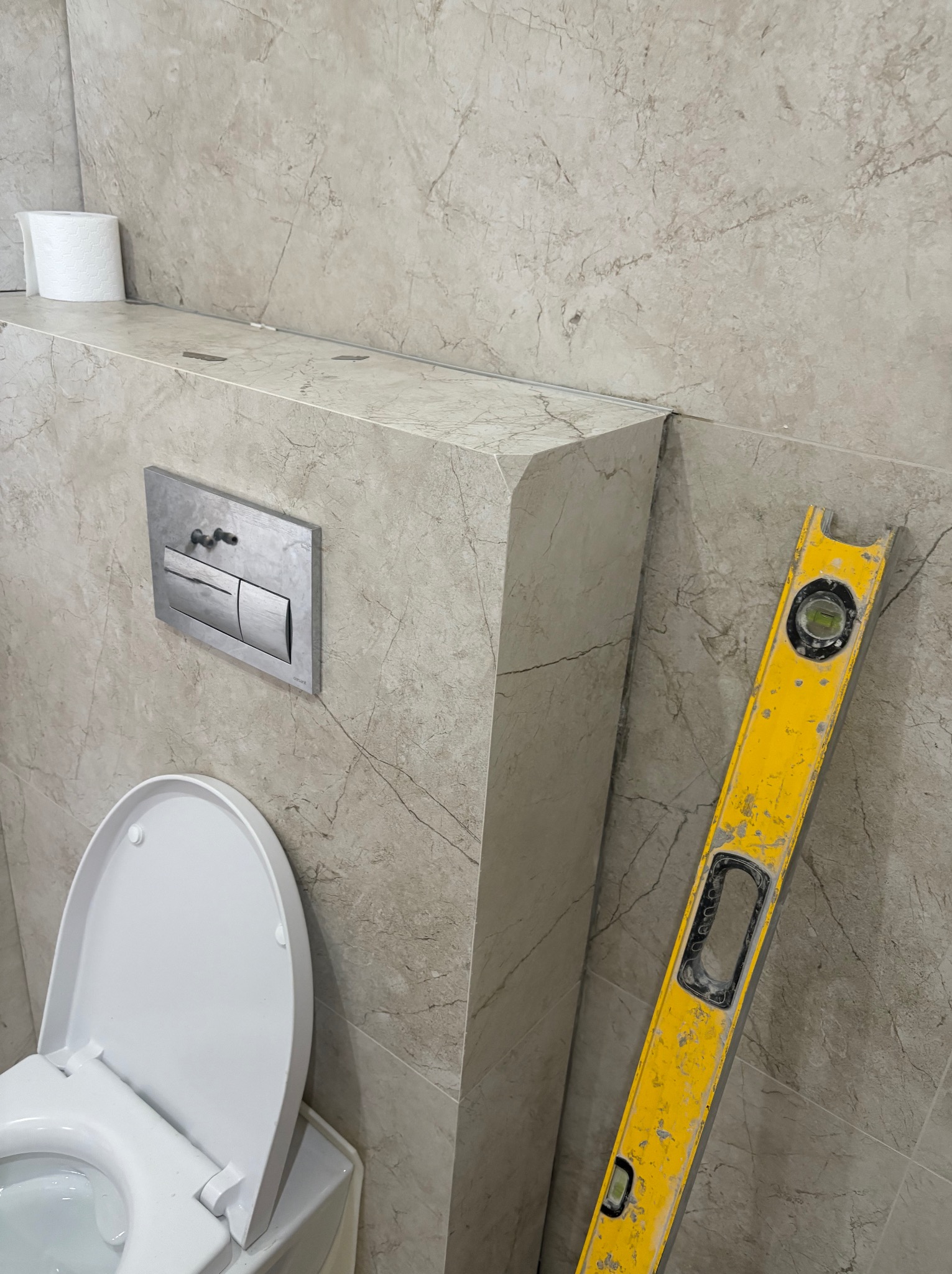 Wykończona toaleta w beżowych płytkach imitujących marmur, z widocznym poziomem wskazującym na precyzję montażu. Uwagę zwraca zabudowa geberitu z półką.