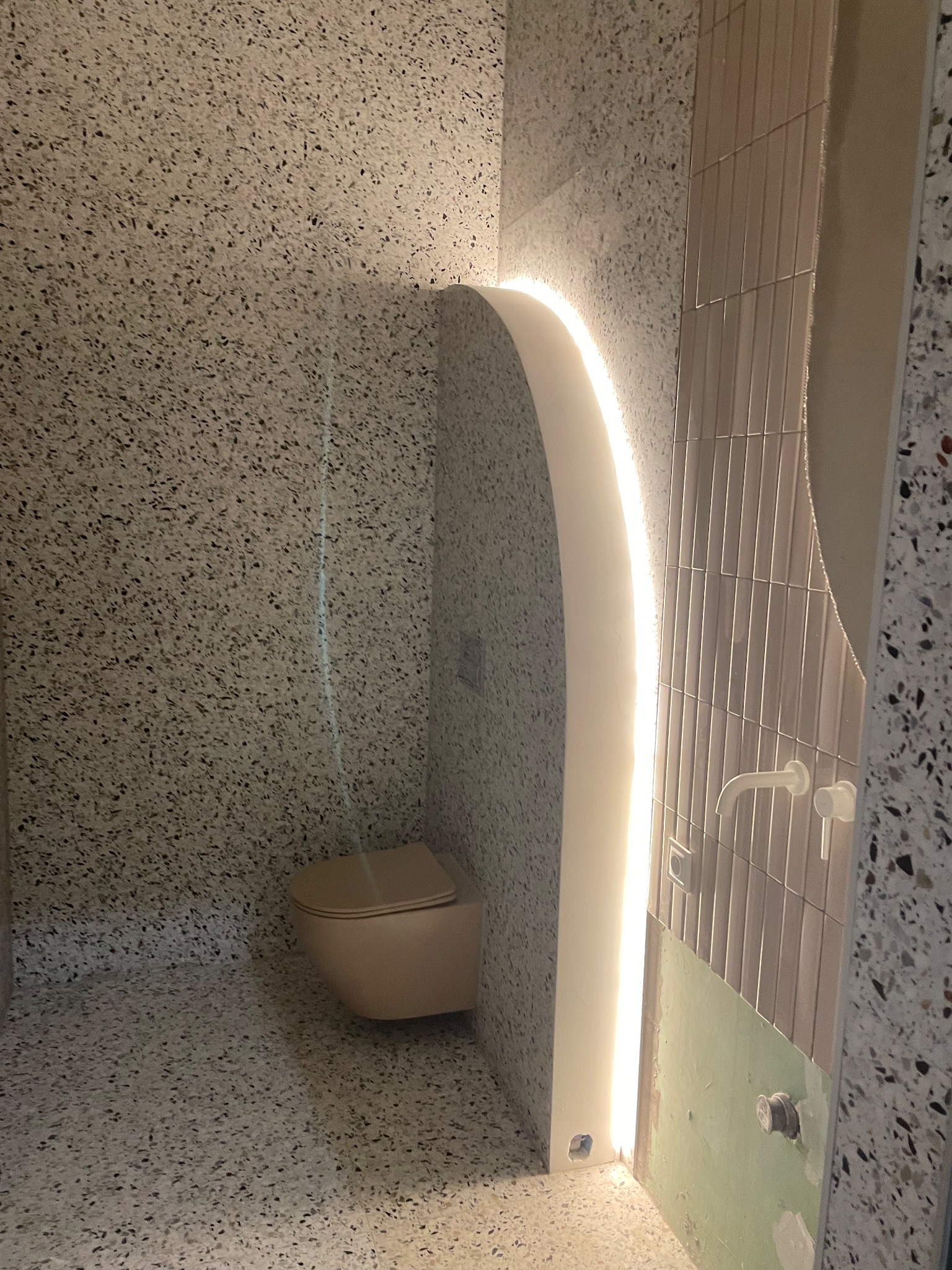 Wykończona łazienka z toaletą w kolorze beżowym, ściany i podłoga wyłożone terazzo, podświetlona wnęka z ledami, pionowe płytki na ścianie.