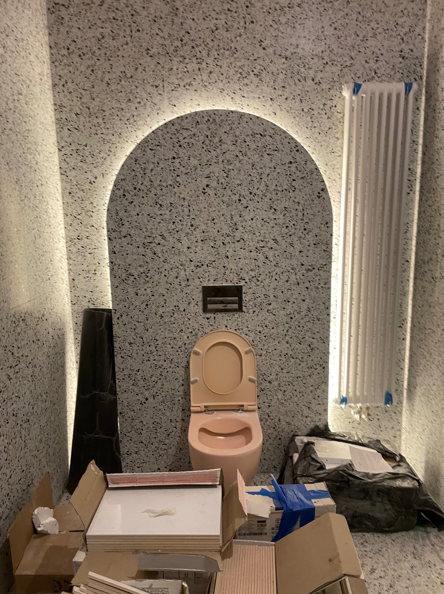 Remont łazienki w trakcie: toaleta w odcieniach różu na tle ściany z lastryko, podświetlona łukiem LED. Widoczne materiały budowlane i grzejnik.