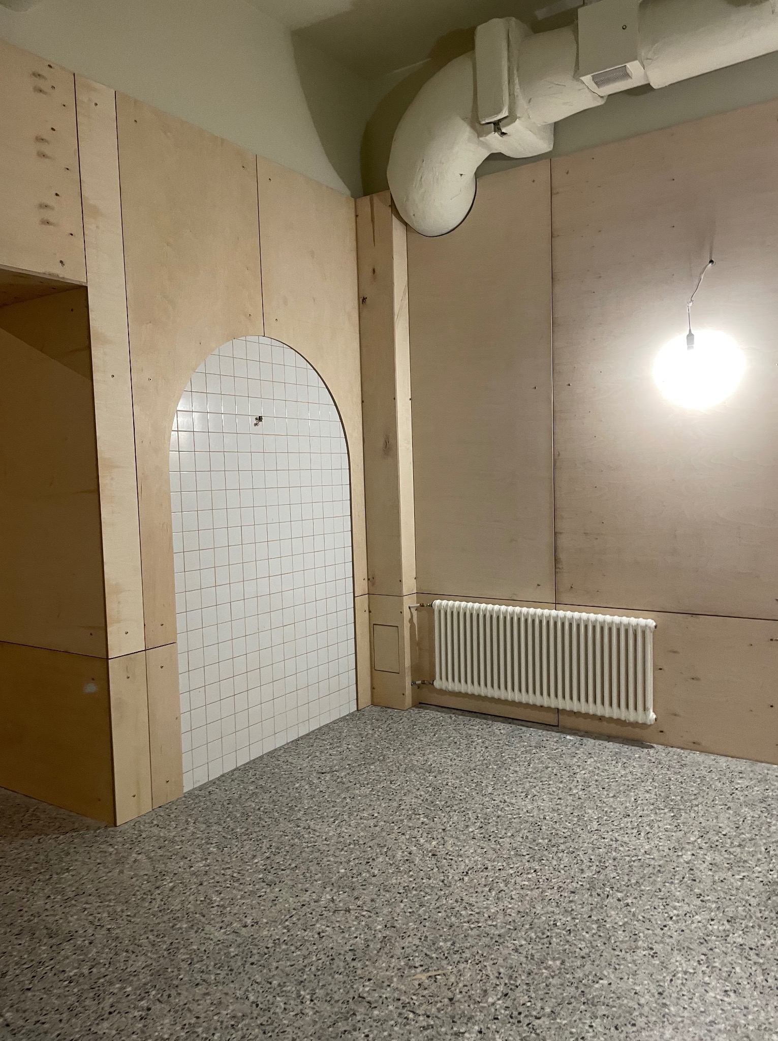 Wnętrze w trakcie wykończenia: surowe ściany z płyt, biała mozaika w łuku, grzejnik żeberkowy, lastryko na podłodze, widoczny kanał wentylacyjny pod sufitem.