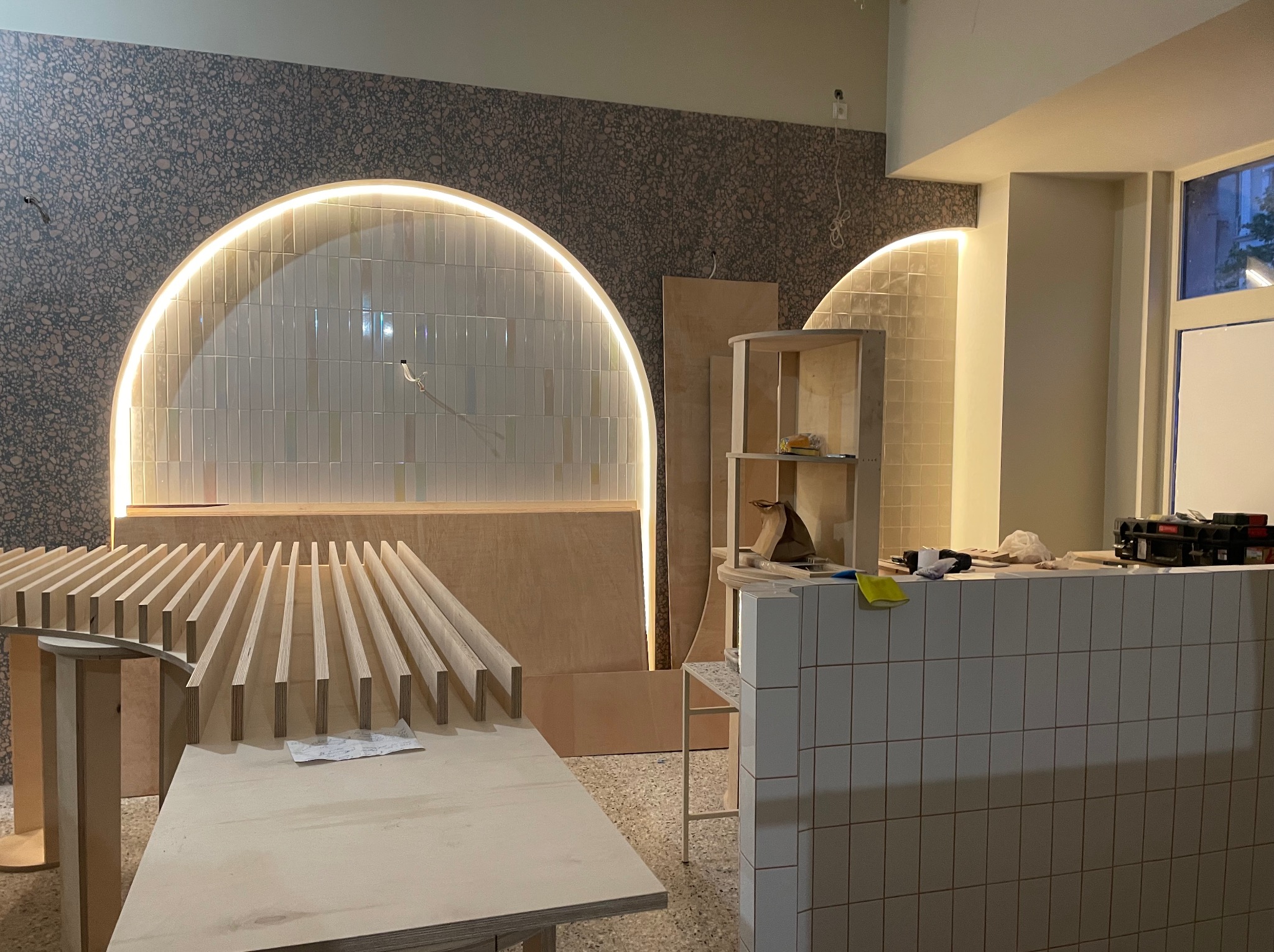 Wnętrze w trakcie wykańczania: drewniane elementy konstrukcyjne, płytki na ścianie i podświetlane łuki. Widoczne narzędzia i materiały budowlane.