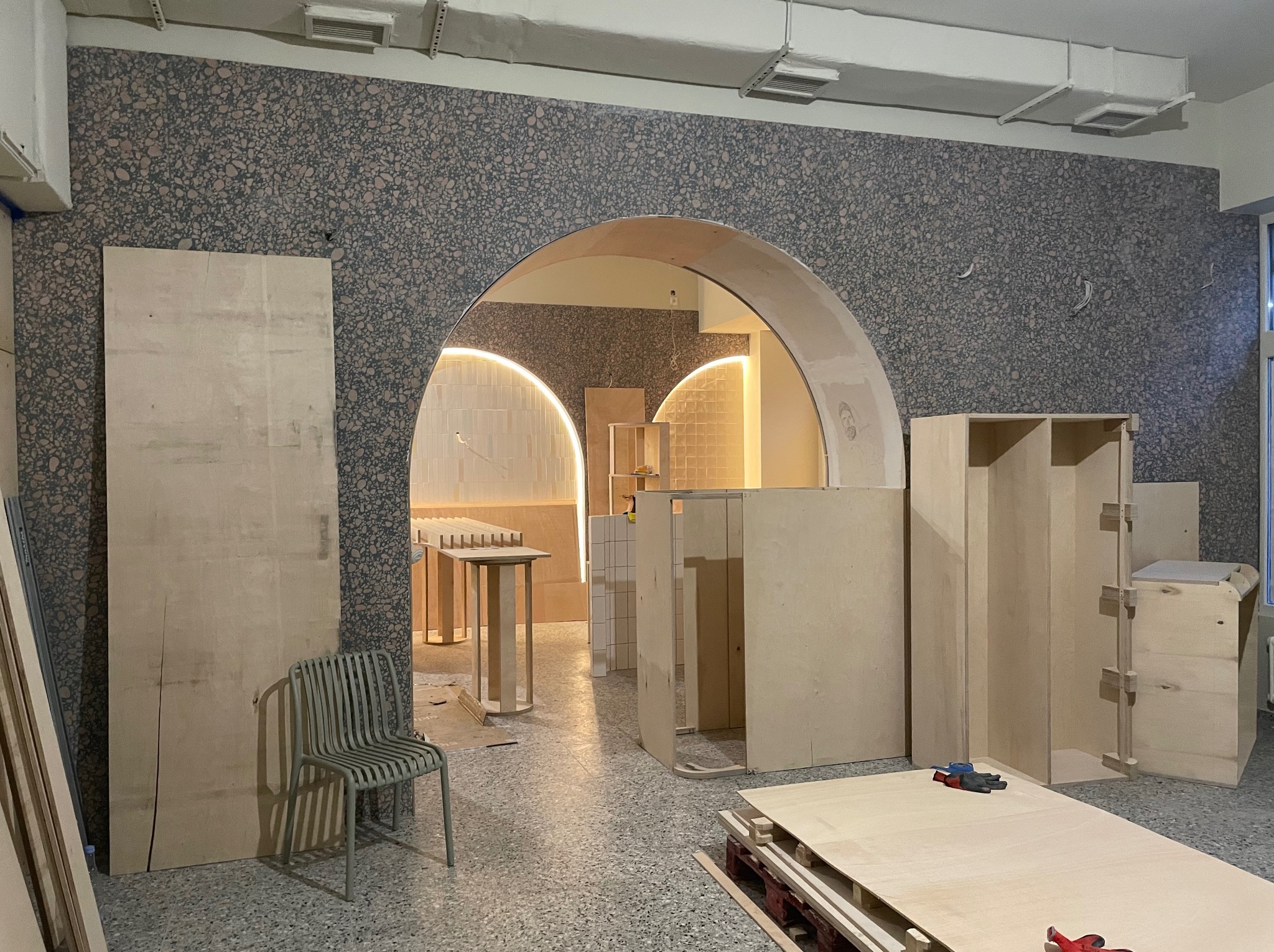 Wnętrze w trakcie wykończenia: surowe drewniane elementy meblowe, łukowate przejście, ściana z dekoracyjnym wzorem, krzesło i narzędzia na podłodze.