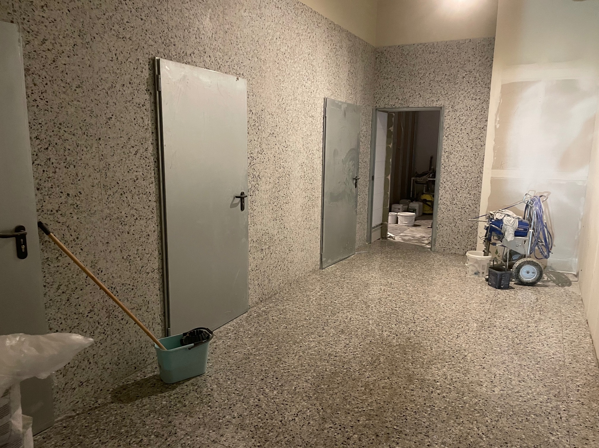 Korytarz po nałożeniu tynku mozaikowego na ścianach i podłodze. Widoczne dwie białe drzwi, wiadro z mopem i agregat malarski.