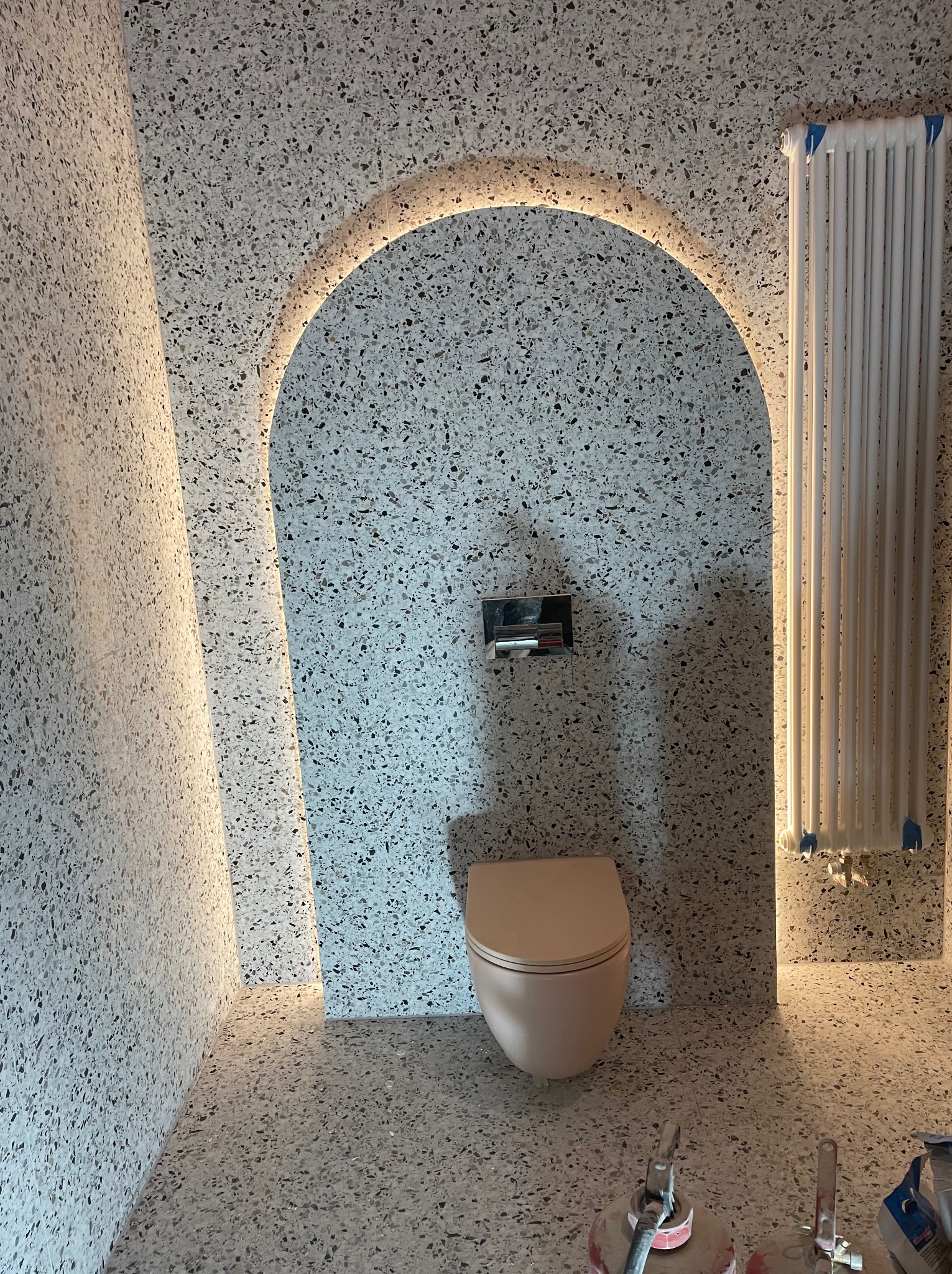 Wykończona łazienka w stylu lastryko z podświetlanym łukiem nad toaletą i grzejnikiem. Widoczne narzędzia sugerują trwające prace wykończeniowe.