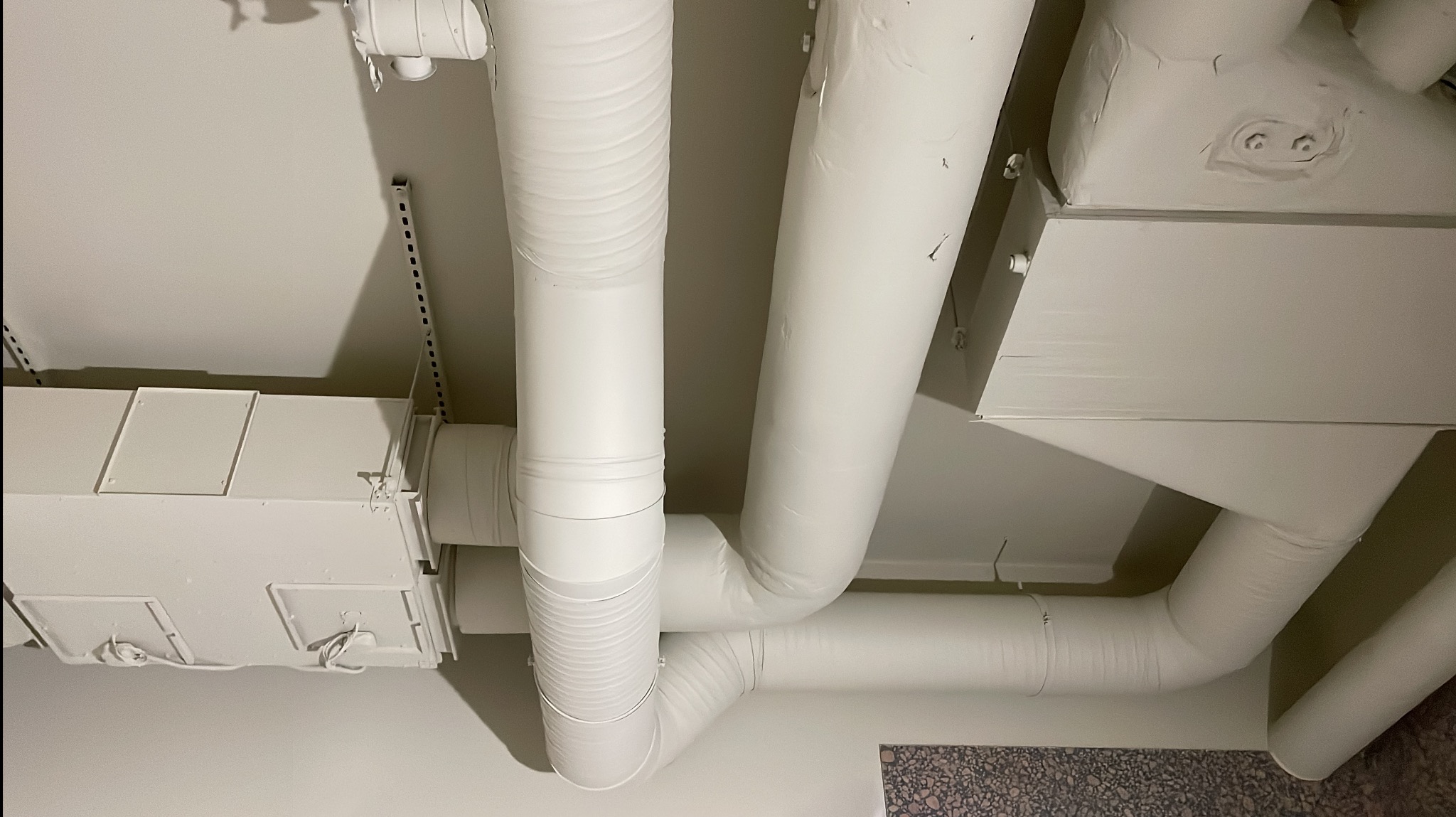 Białe rury wentylacyjne i klimatyzacyjne pod sufitem, widok z dołu. Elementy instalacji pomalowane na biało, widoczne łączenia i mocowania.