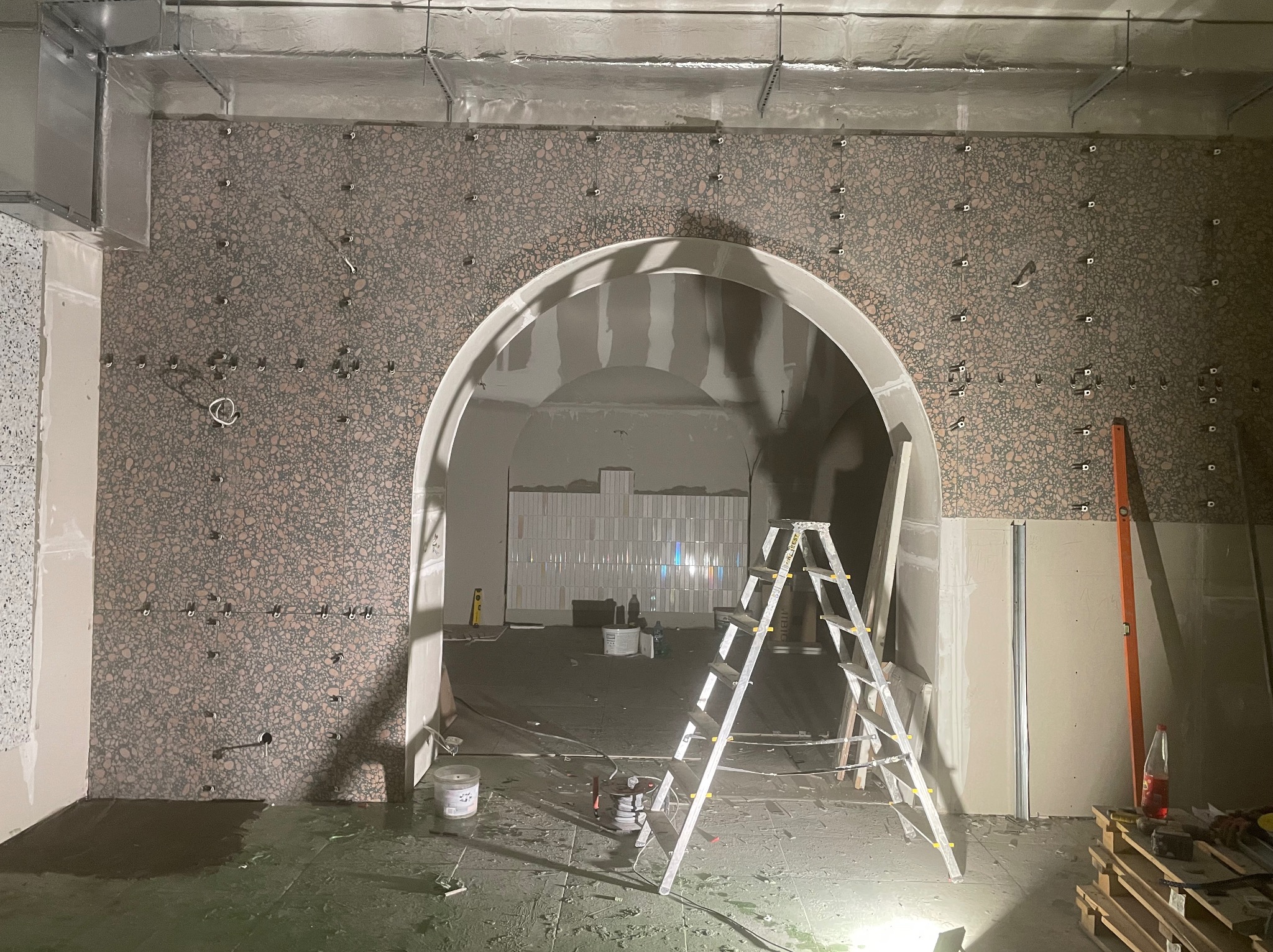 Montaż płytek imitujących lastryko na ścianie z łukowym przejściem. Widoczna drabina, poziomica i narzędzia. Prace wykończeniowe w trakcie realizacji.
