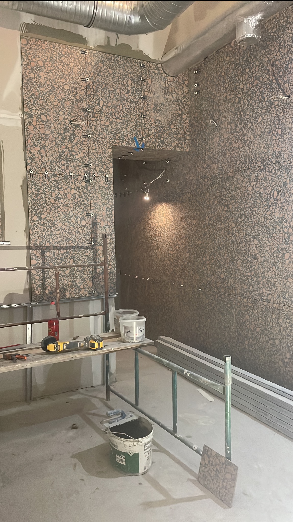 Montaż płytek o wzorze lastryko na ścianie z użyciem poziomujących klipsów. Widoczne narzędzia i materiały na stole roboczym. Prace w toku, surowy stan.