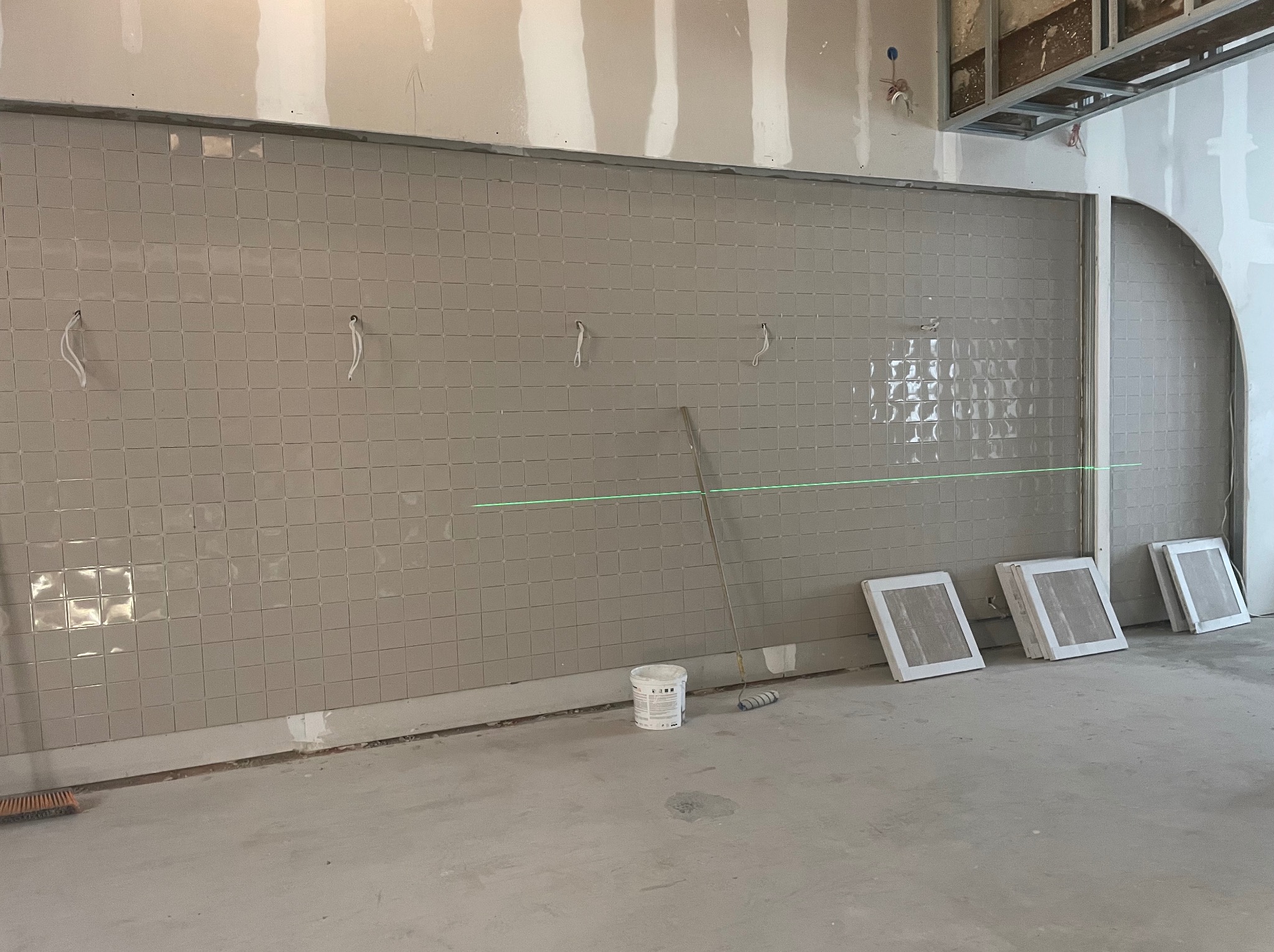 Ściana z szarych, kwadratowych płytek, wyznaczona poziomą linią lasera. Na podłodze wiadro z farbą, wałek i zapasowe płytki. Widoczne kable i surowy stan wykończenia.