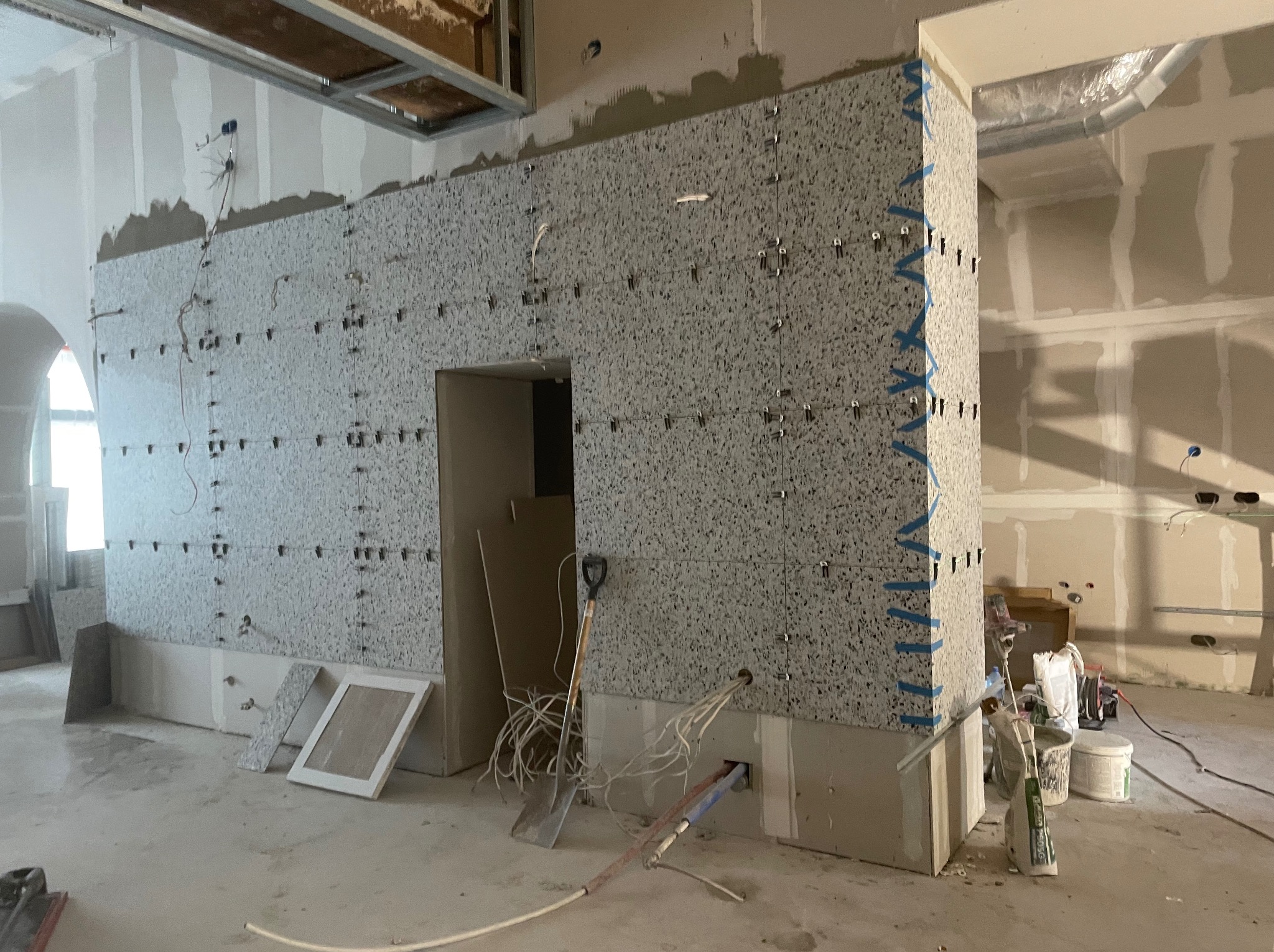 Ściana z szarych płytek w trakcie montażu, z widocznymi krzyżykami dystansowymi i taśmą maskującą na narożniku. Przewody i narzędzia na podłodze.