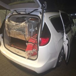 Wszystko co wejdzie na pakę - Biały minivan z otwartym bagażnikiem, w którym znajduje się mebel owinięty folią i czerwony koc. Częściowo widoczna przyczepka z plandeką.