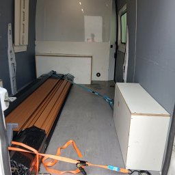 Wszystko co wejdzie na pakę - Wnętrze busa transportowego z szarymi ścianami, białymi panelami ochronnymi i widocznym odbiciem osoby. Długie, brązowe elementy i biała skrzynia zabezpieczone pomarańczowymi pasami.