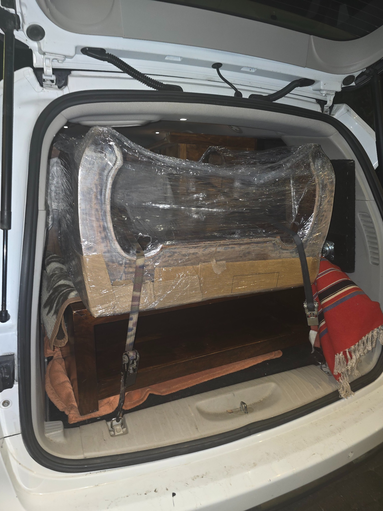 Mebel zabezpieczony folią i pasami w bagażniku białego samochodu. Koce i pledy dopełniają ochronę podczas transportu.
