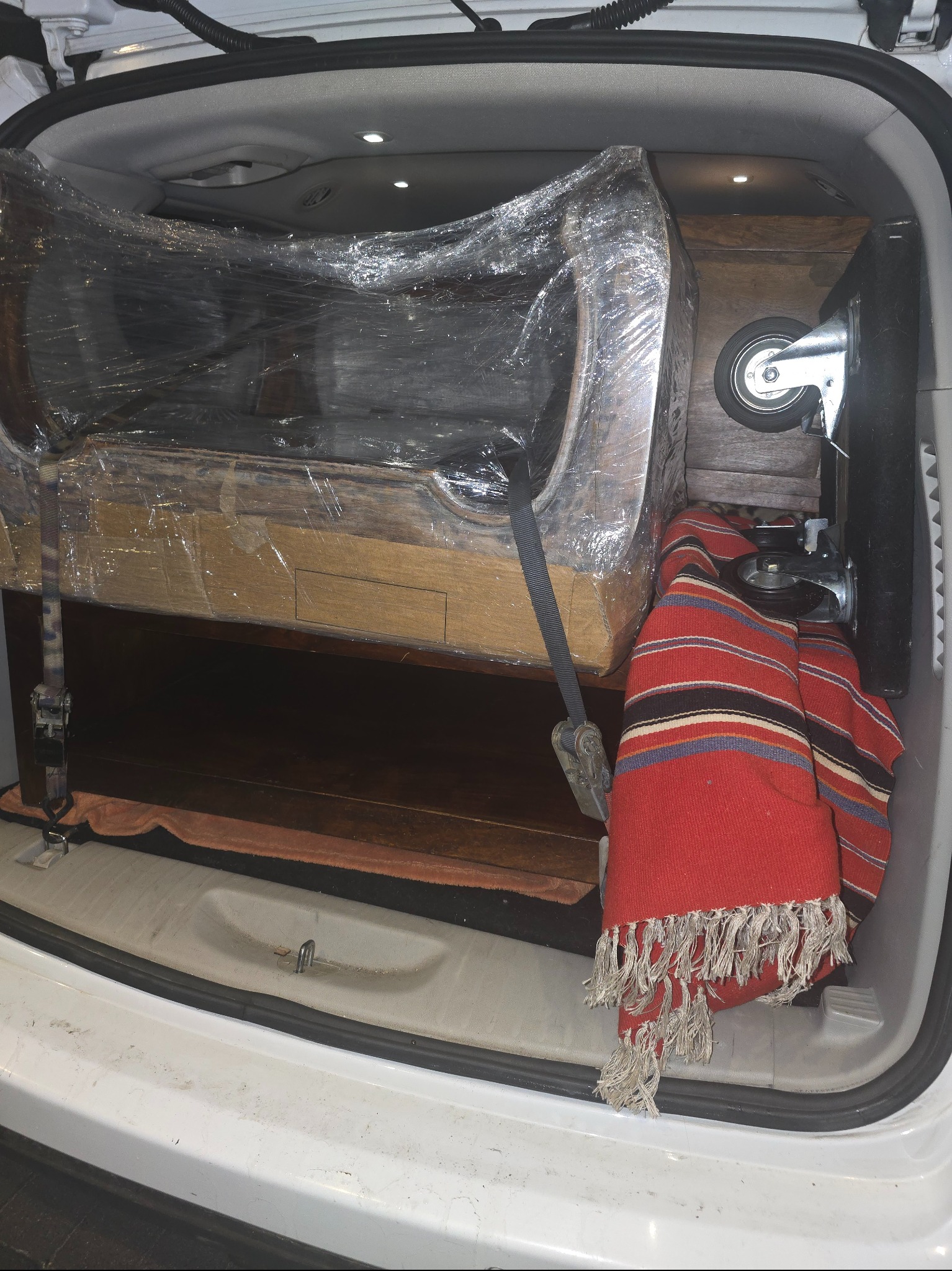 Pianino zabezpieczone folią i pasami w bagażniku białego auta, gotowe do transportu. Czerwony pled z frędzlami dodaje koloru. Widoczne kółka transportowe.