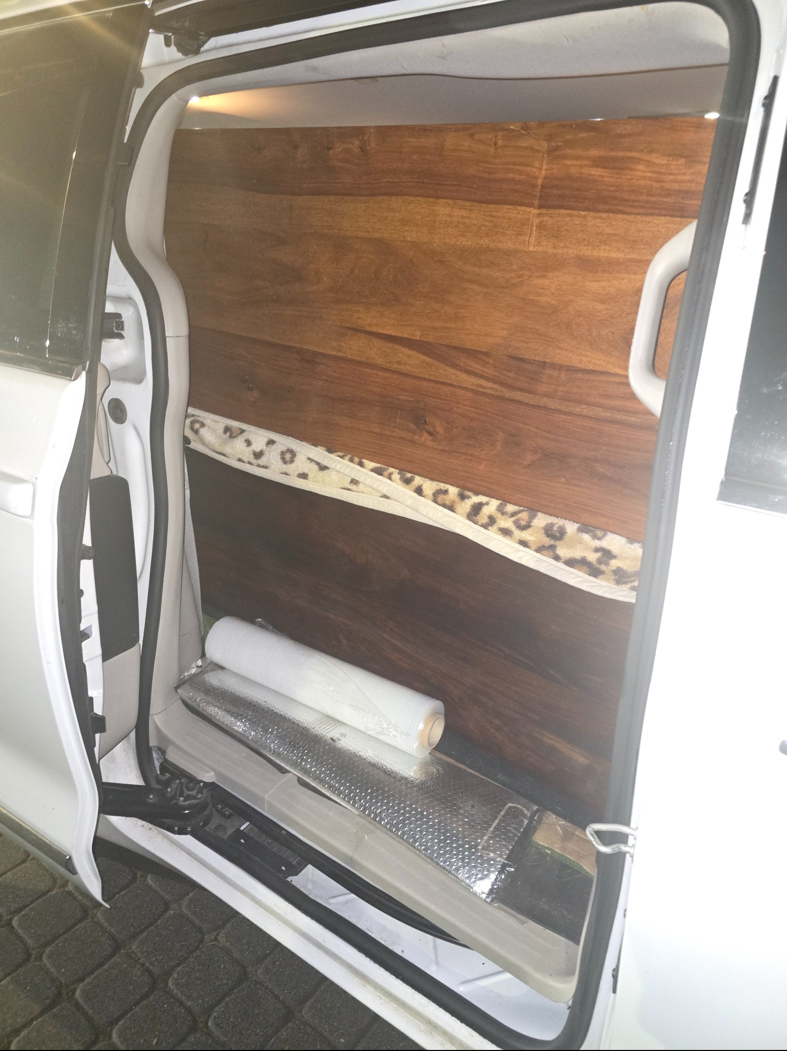 Wnętrze białego busa z drewnianą zabudową, zabezpieczone folią i materiałem w panterkę, gotowe do transportu. Widok z otwartych drzwi.