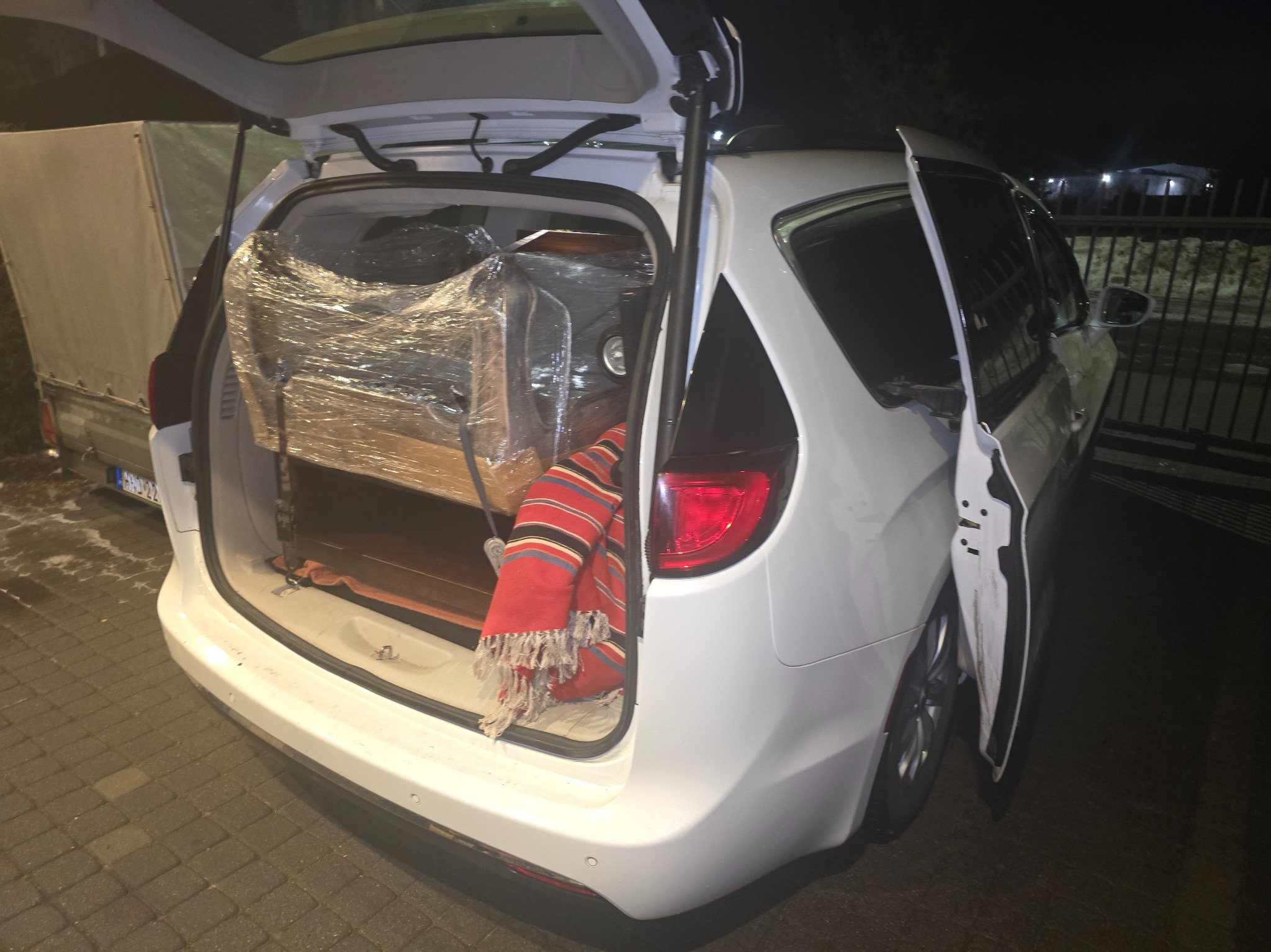 Biały minivan z otwartym bagażnikiem, w którym znajduje się mebel owinięty folią i czerwony koc. Częściowo widoczna przyczepka z plandeką.