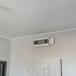 USŁUGI U RAFAŁA Rafał Zieliński - Białe drzwi z panelami i szara ściana z zamontowaną skrzynką elektryczną w jasnym wnętrzu. Minimalistyczny design, neutralna kolorystyka.