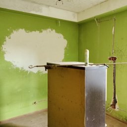 USŁUGI U RAFAŁA Rafał Zieliński - Pokój w trakcie remontu: zielone ściany z fragmentami szpachli, widoczne instalacje elektryczne, prowizoryczne oświetlenie i mebel z napisem 'GORA'.