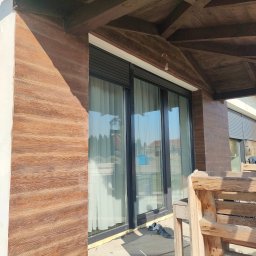 USŁUGI U RAFAŁA Rafał Zieliński - Elewacja domu z drewnopodobną okładziną, widoczne przesuwne okna z roletami zewnętrznymi i drewniana konstrukcja zadaszenia tarasu.